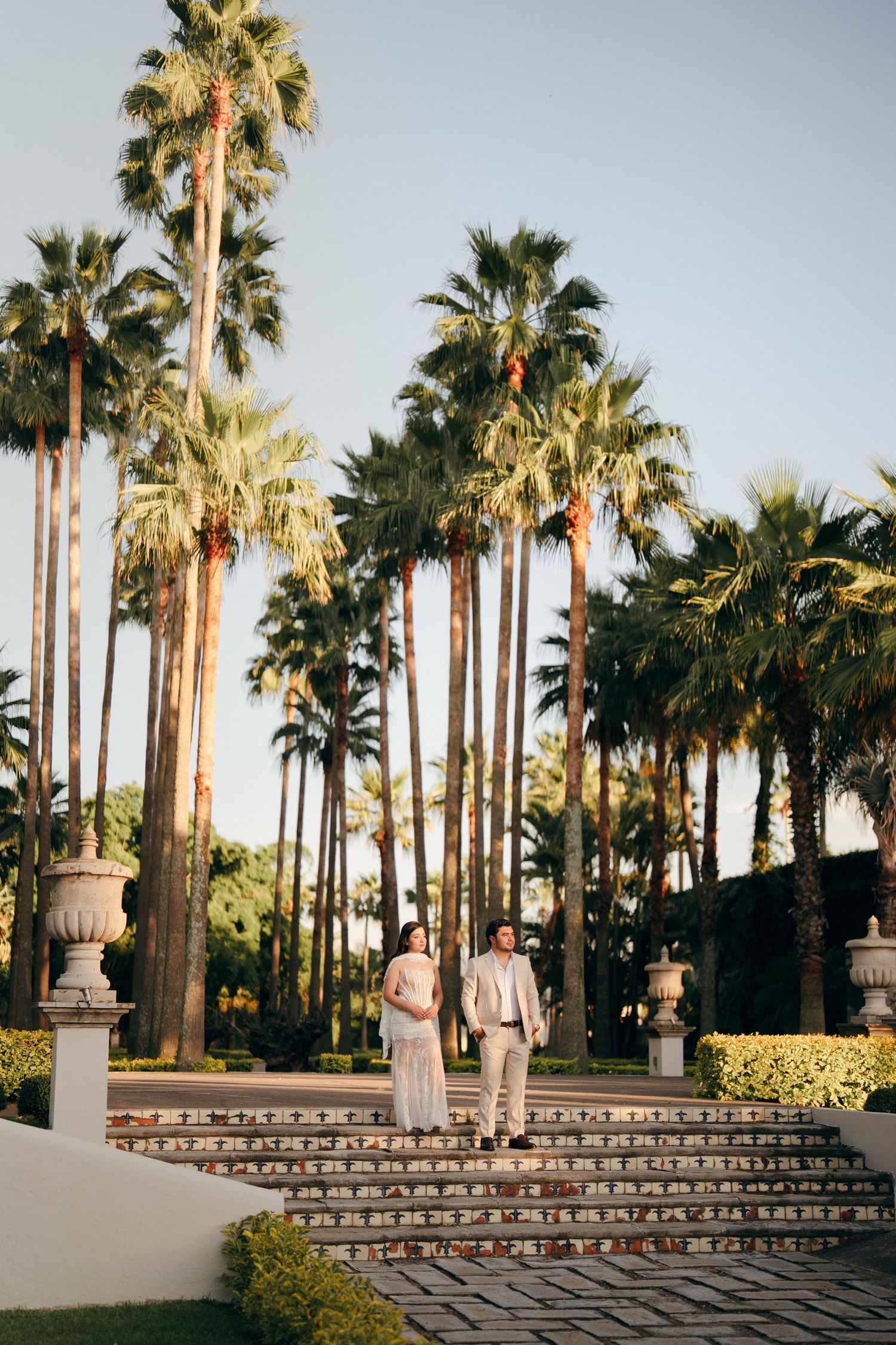 Bride and groom walk down stone steps surrounded by tall palm trees at a sunny outdoor wedding venue.