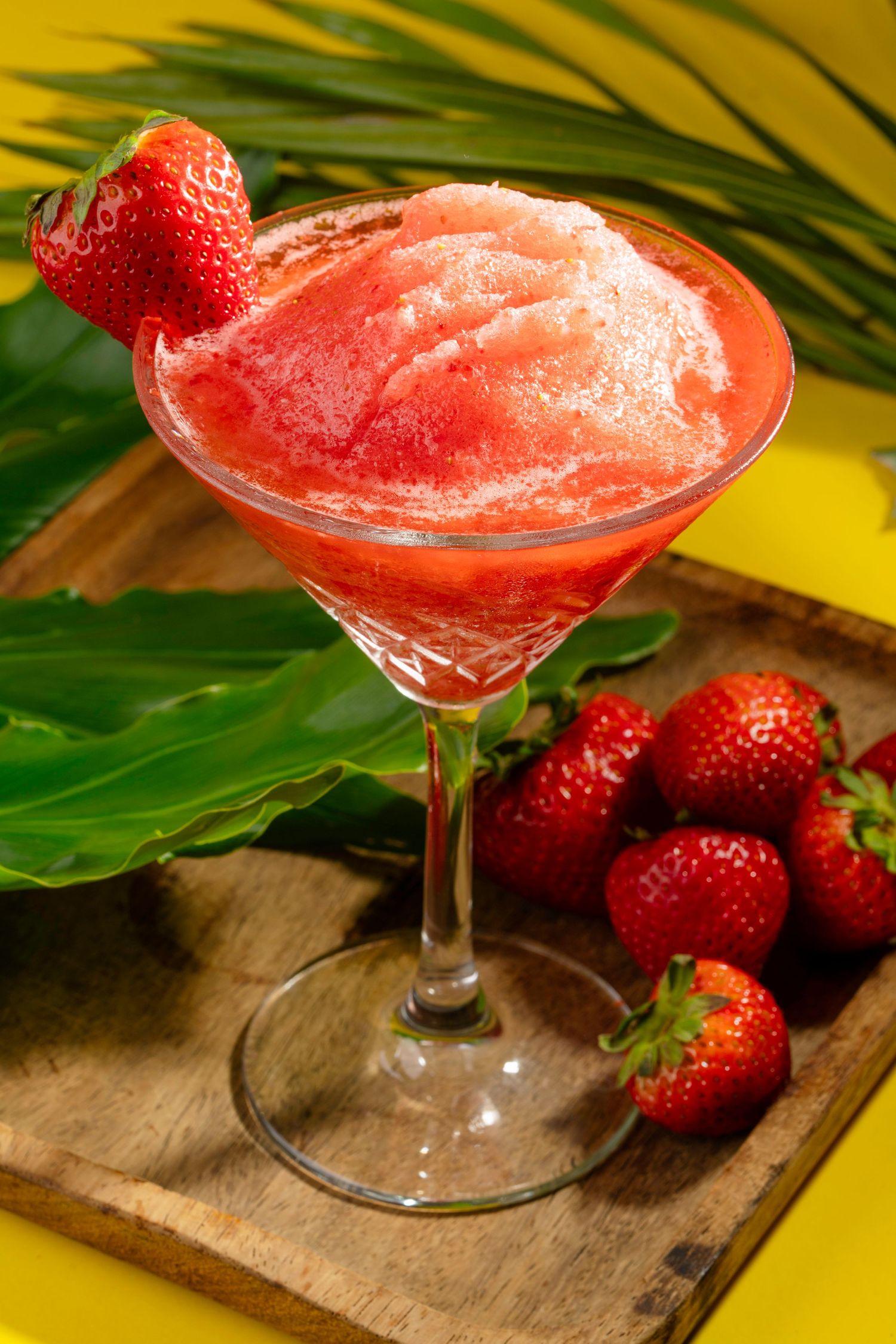 Frozen strawberry margarita in cocktail glass with fresh strawberries and tropical decor.