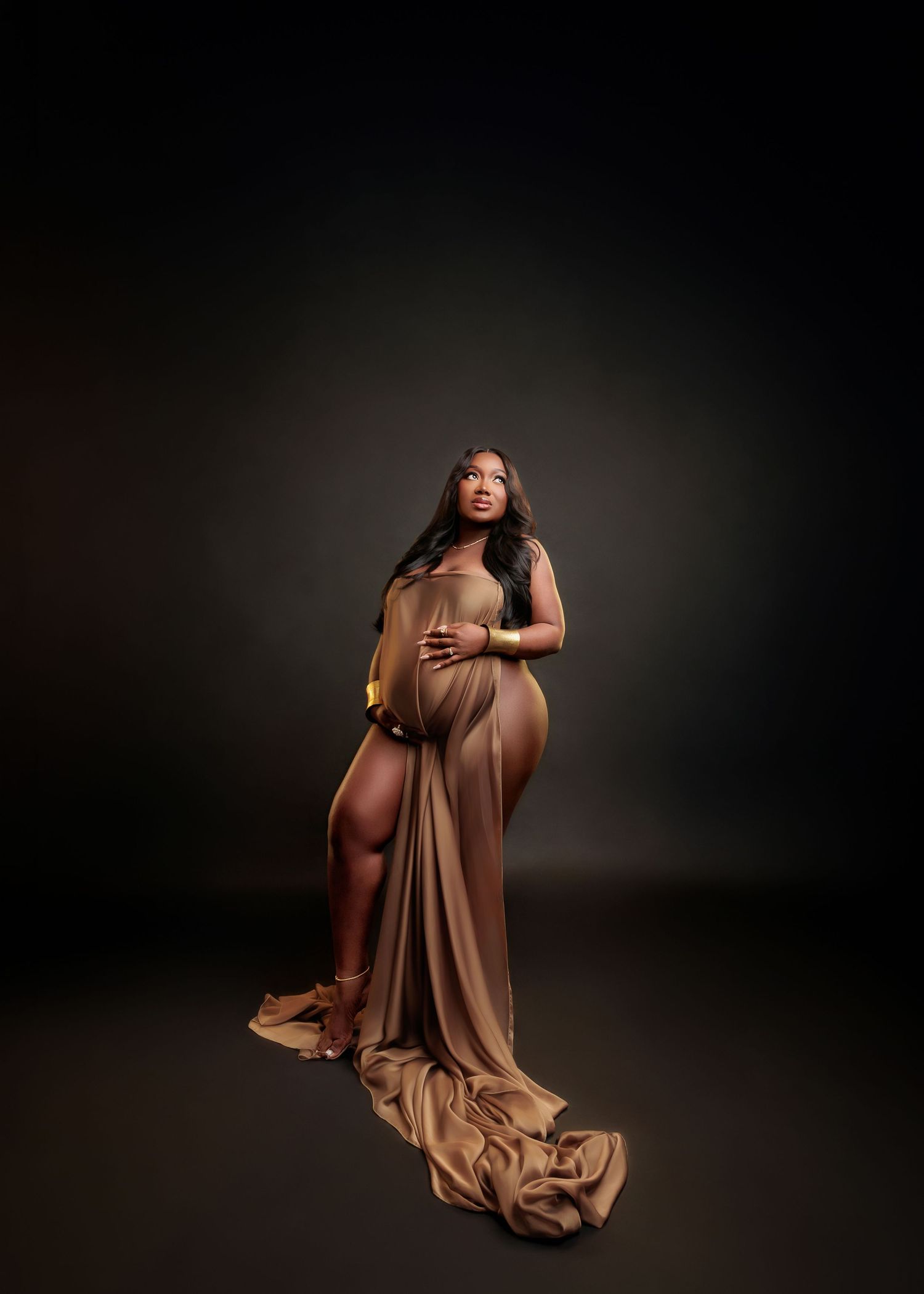 Pregnant woman in elegant golden draped gown posing gracefully in a professional maternity photo shoot against dark backdrop.