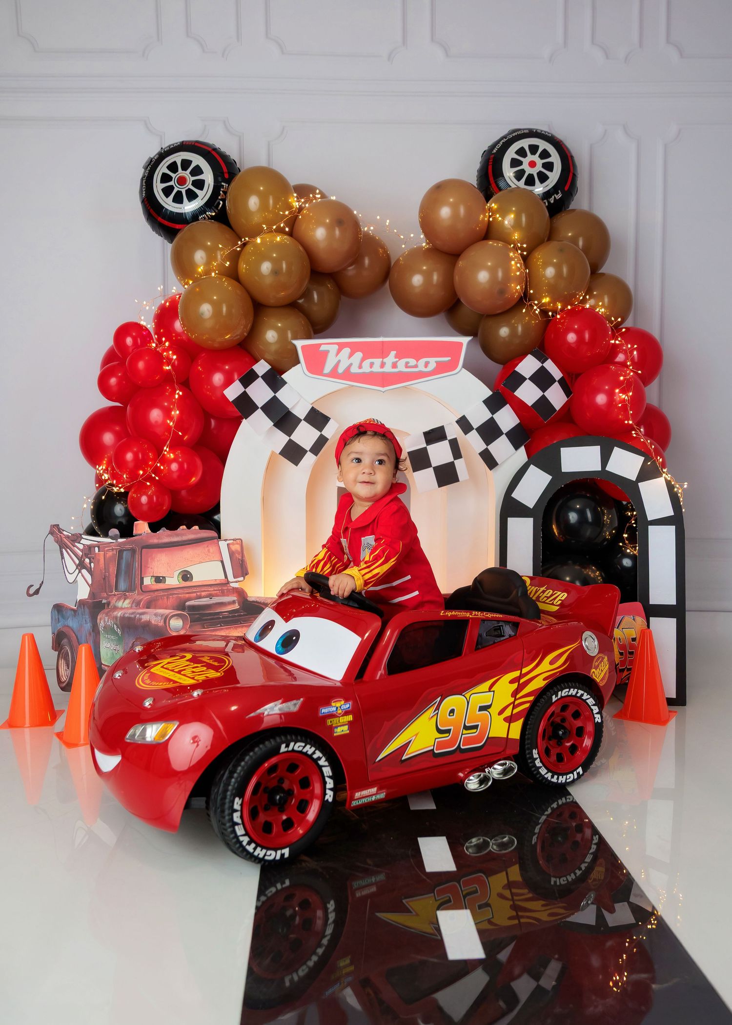 Toddler boy in Lightning McQueen costume sitting in toy car at Cars themed birthday setup.