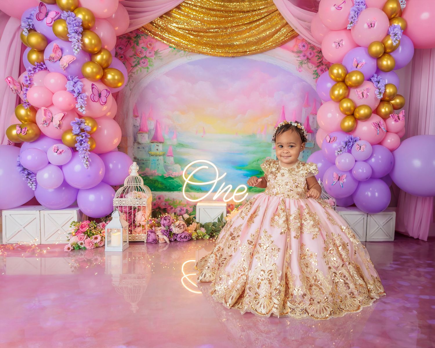Young girl in elegant gold floral gown at princess themed first birthday with pink purple balloons.