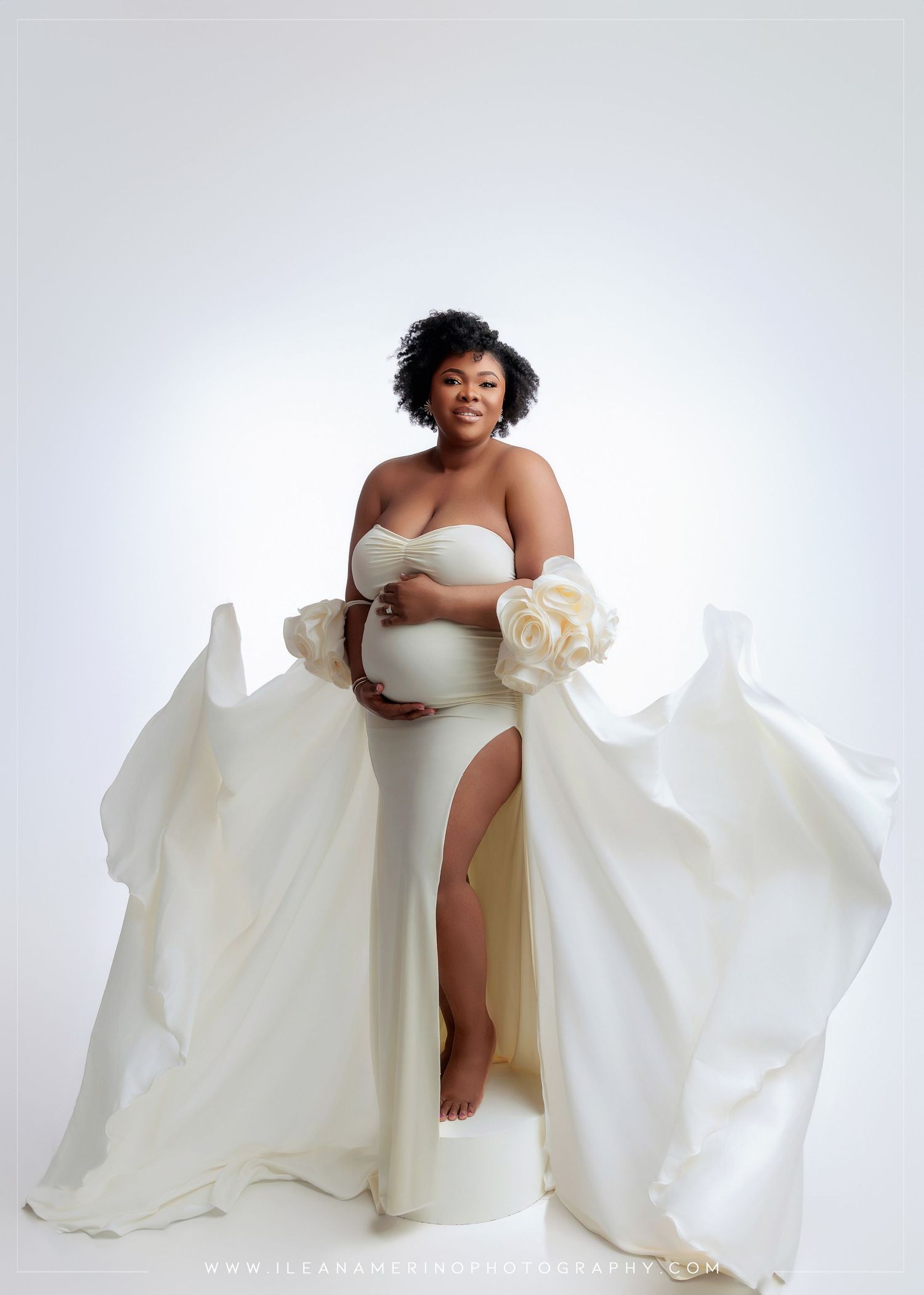 Pregnant woman in white strapless gown with floral sleeves poses gracefully in studio.