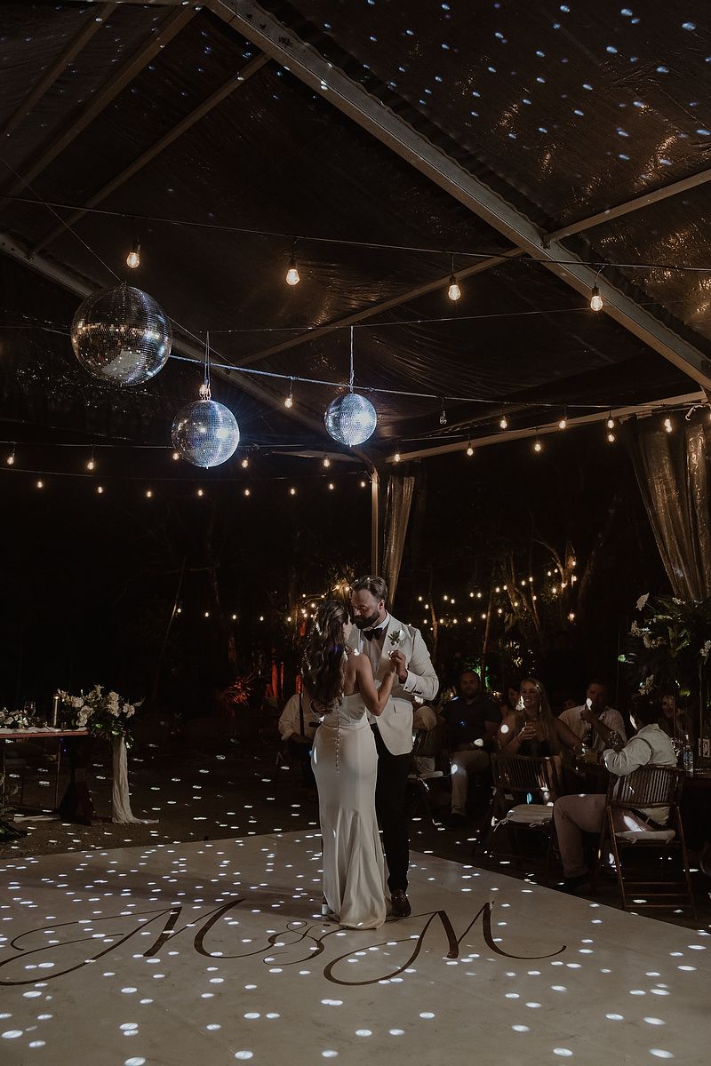 Couple shares first dance under disco balls and string lights on monogrammed dance floor at elegant outdoor wedding reception.