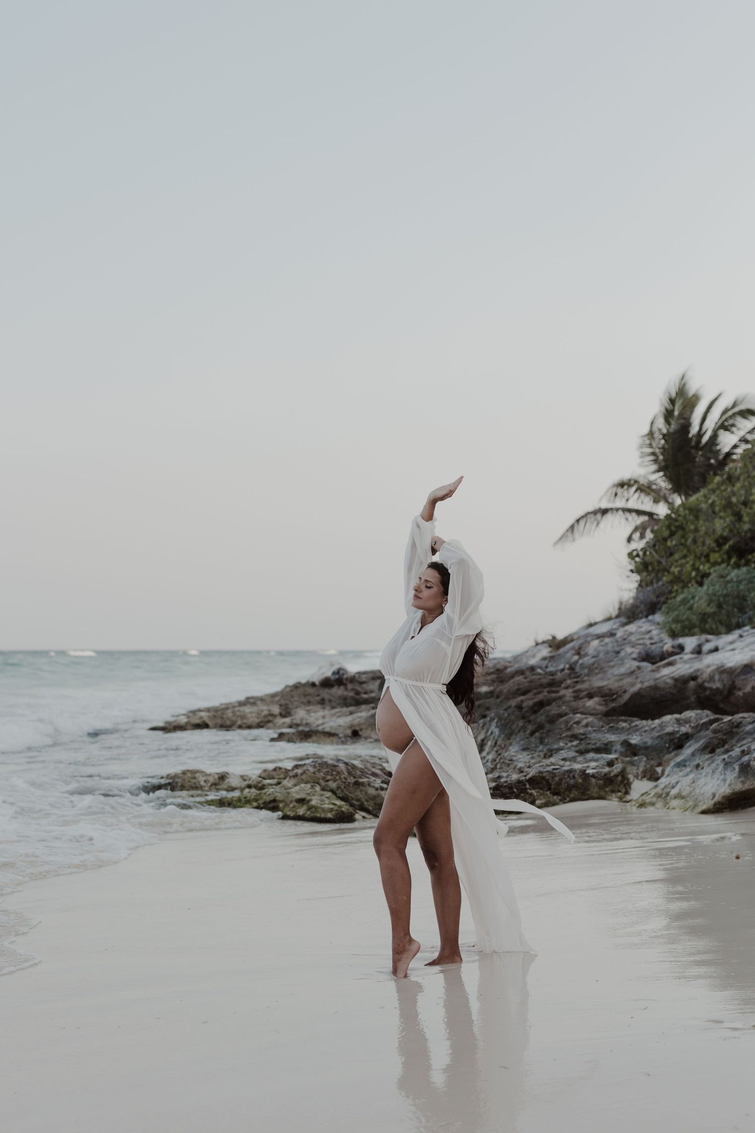 Graceful dancer in white poses on a tropical beach with rocks and palm trees Portrait session at Our Habitas Tulum.