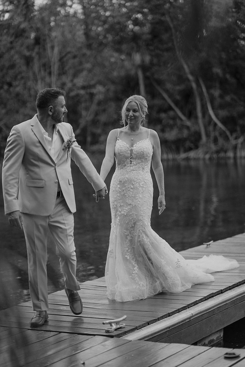 Black and white photo of a bride and groom holding hands on a wooden dock by a lake on their wedding day.