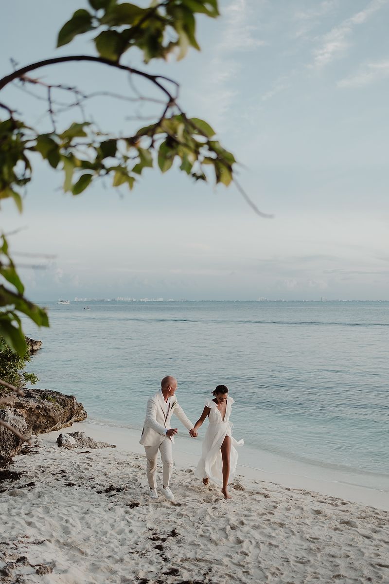 Couple in white wedding attire walking hand in hand on a tropical white sand beach with calm turquoise ocean waters.