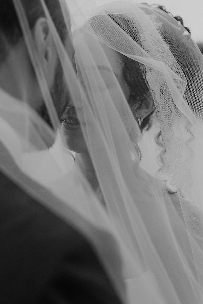 Black and white photo of bride and groom sharing an intimate moment under a flowing wedding veil.