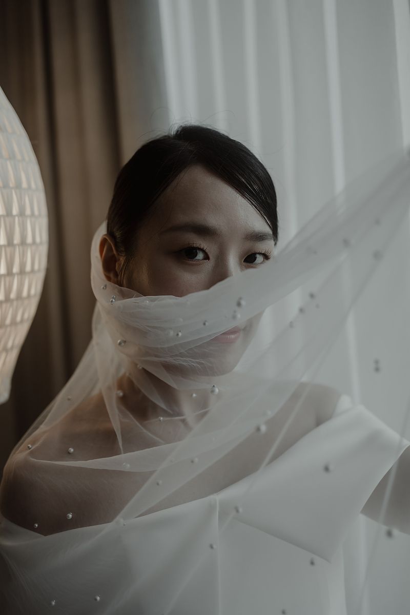 A bride peers over a pearl-embellished white veil near a window, her dark hair pulled back elegantly.