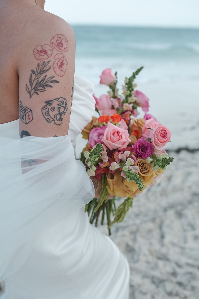 Tattooed bride in white gown holds colorful pink and yellow floral bouquet on a beach.