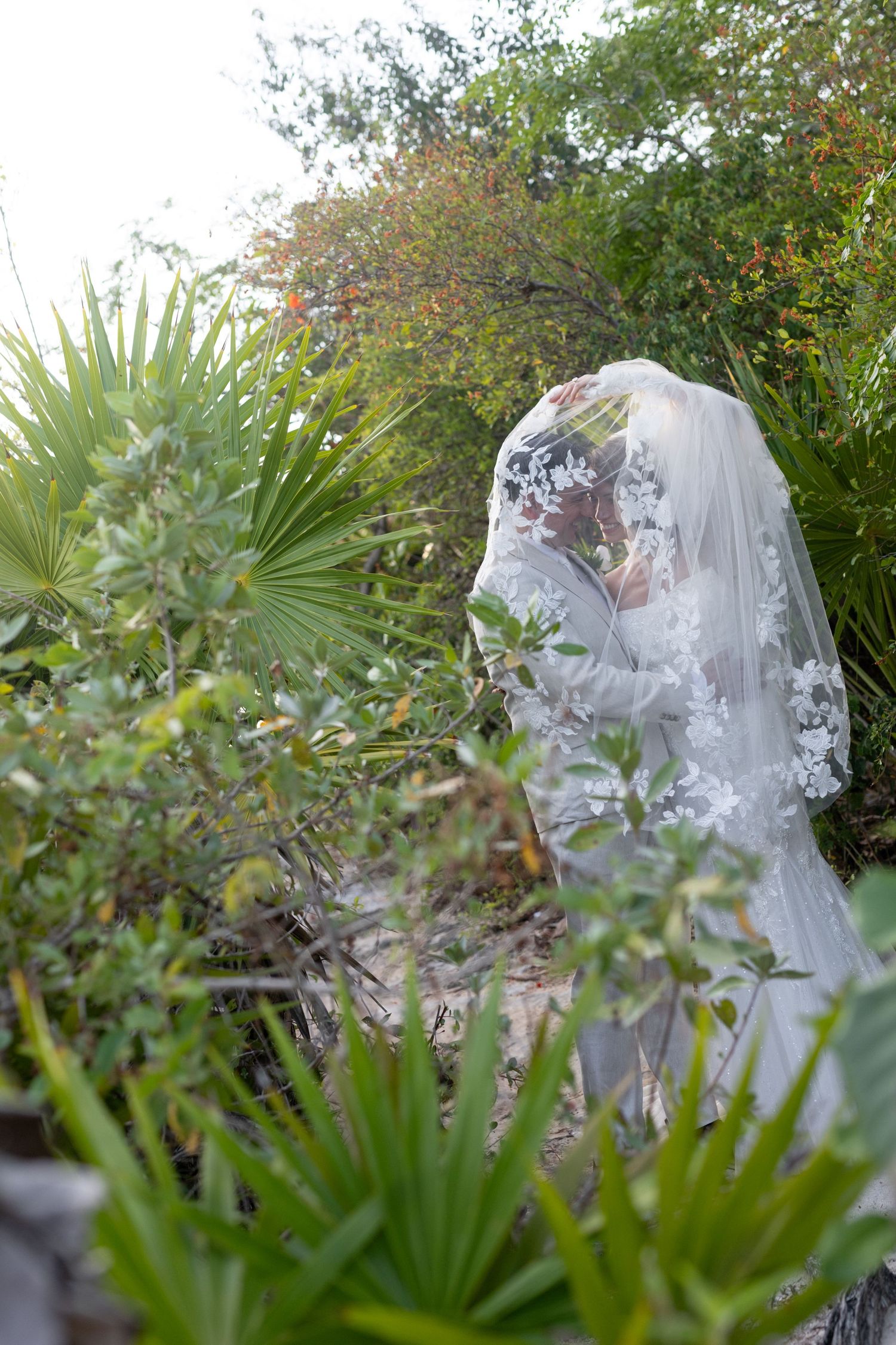 Bride in lace veil surrounded by lush tropical greenery and palm trees in an outdoor garden wedding setting.