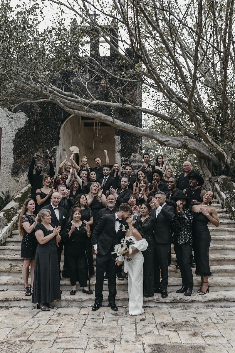 Wedding couple kissing on stone steps surrounded by guests in black attire at an outdoor venue with rustic stone arch.