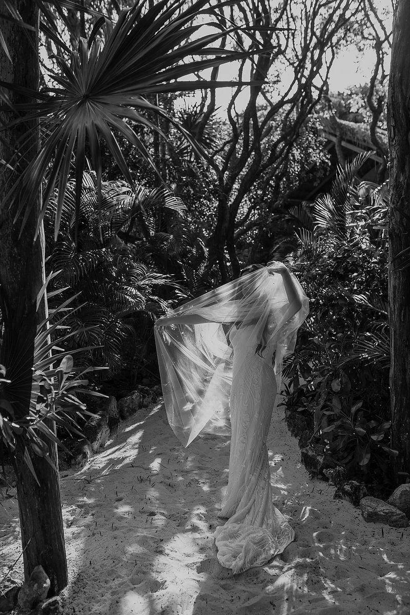 Black and white photo of a person in a flowing white dress on a sandy path surrounded by tropical trees.