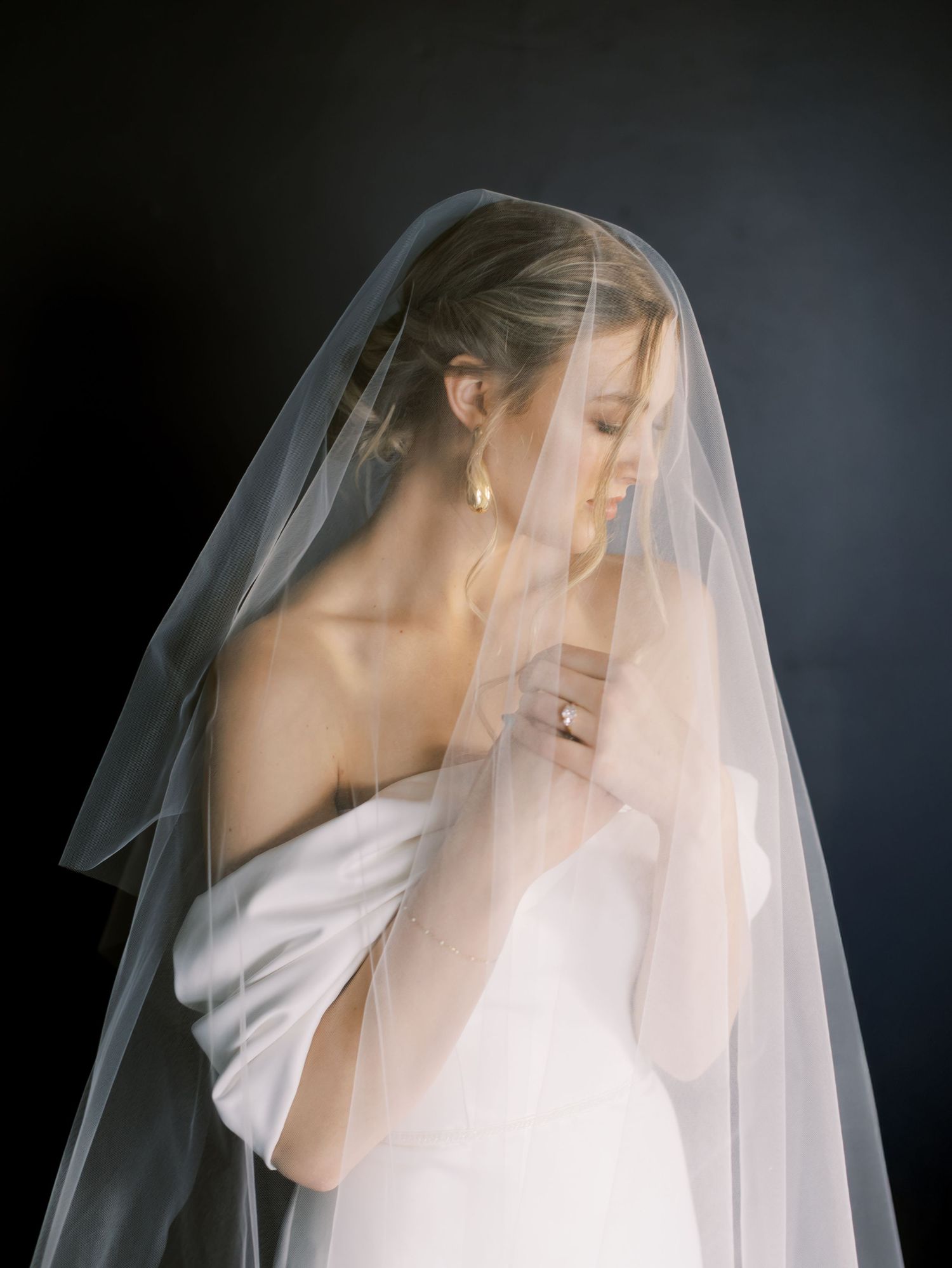 Ethereal bridal portrait with flowing sheer veil and off-shoulder white gown against dark background.