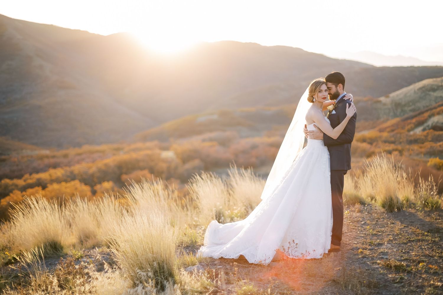 Bride and groom embracing at golden sunset on mountain hilltop, wedding dress veil flowing, warm autumn landscape backdrop.