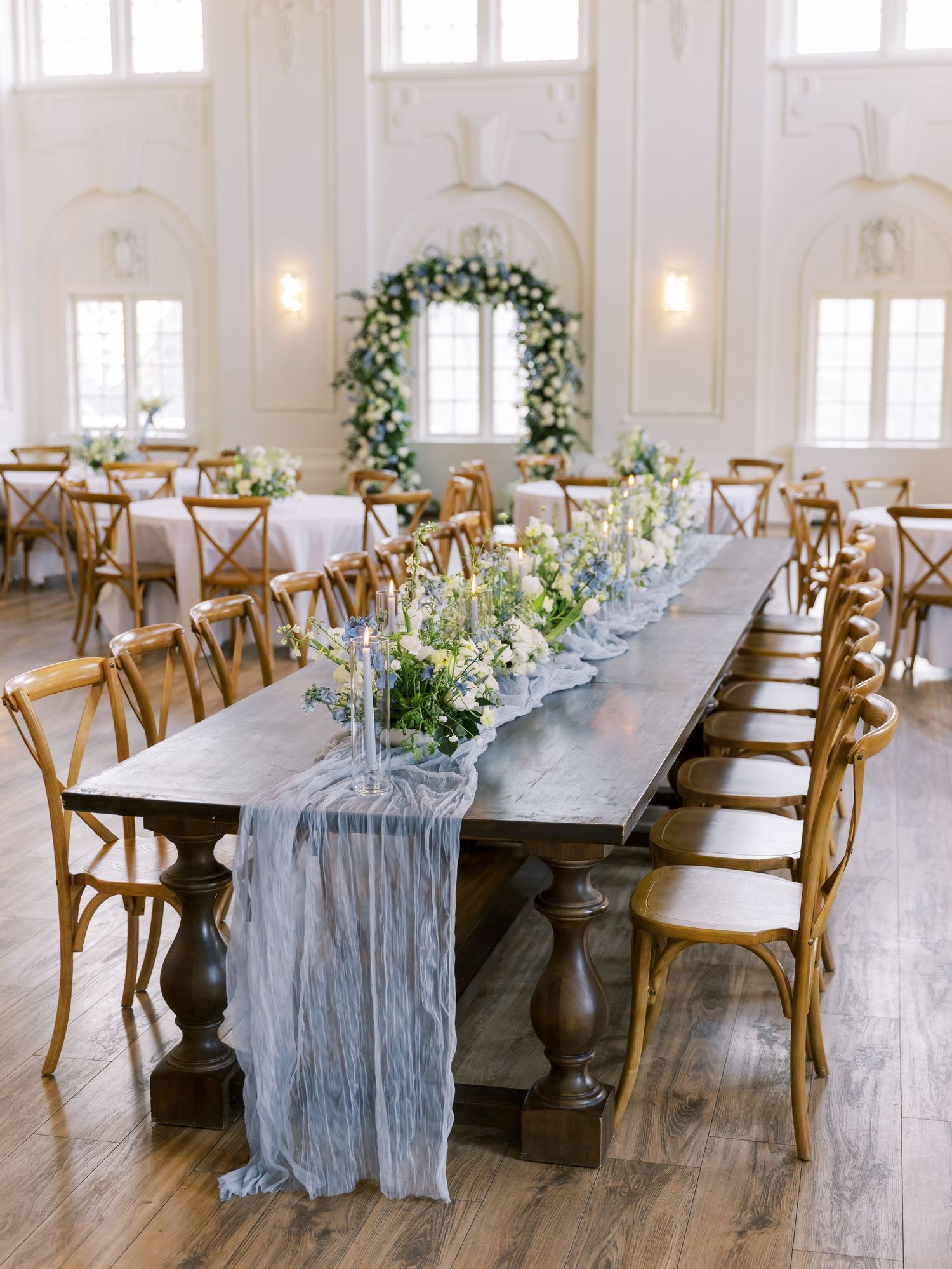 Elegant wedding reception tables with light blue gauze runners and wooden cross-back chairs in a bright white venue.