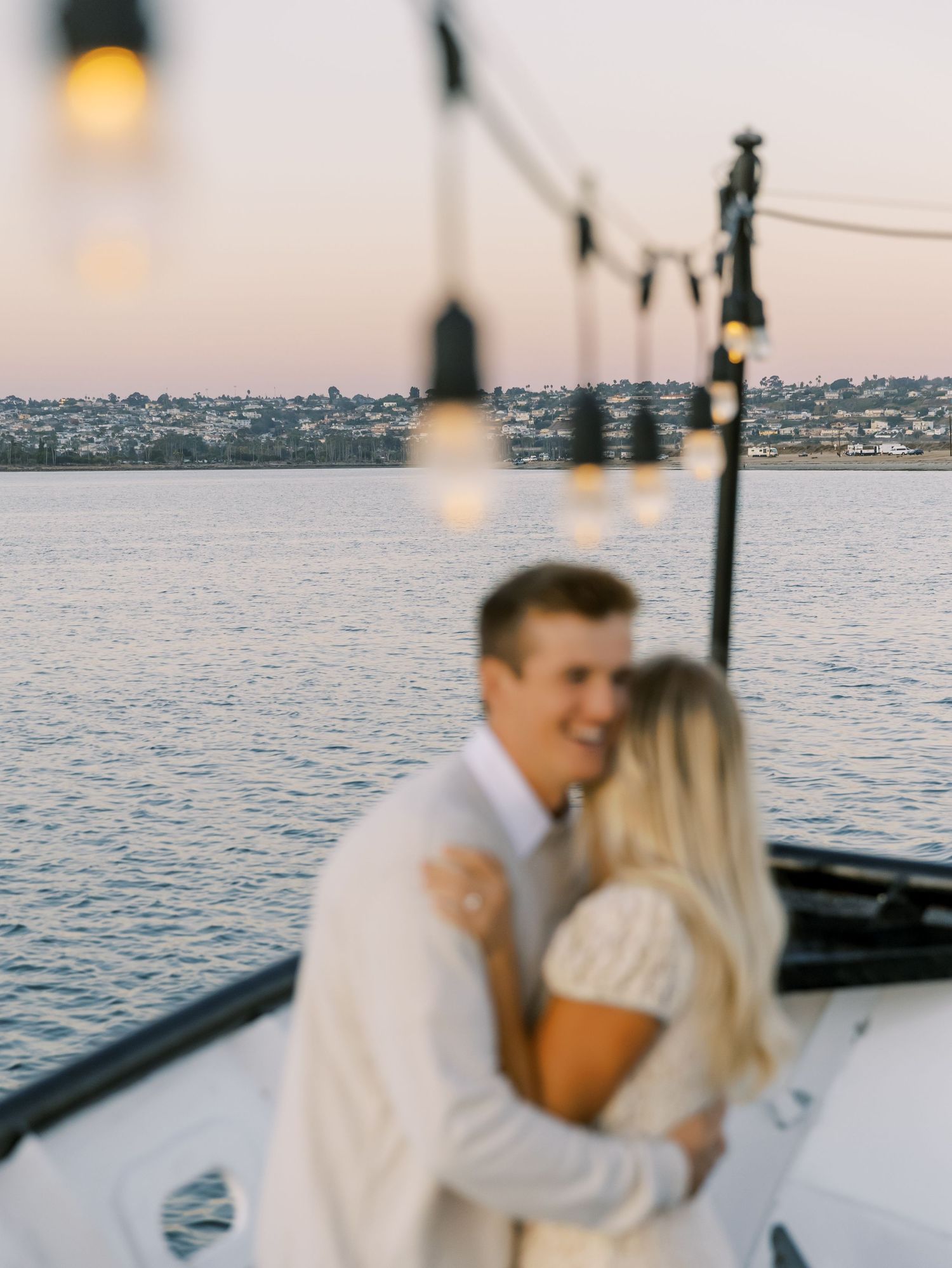 A couple shares a romantic moment on a boat at sunset with string lights glowing overhead and the ocean in the background.