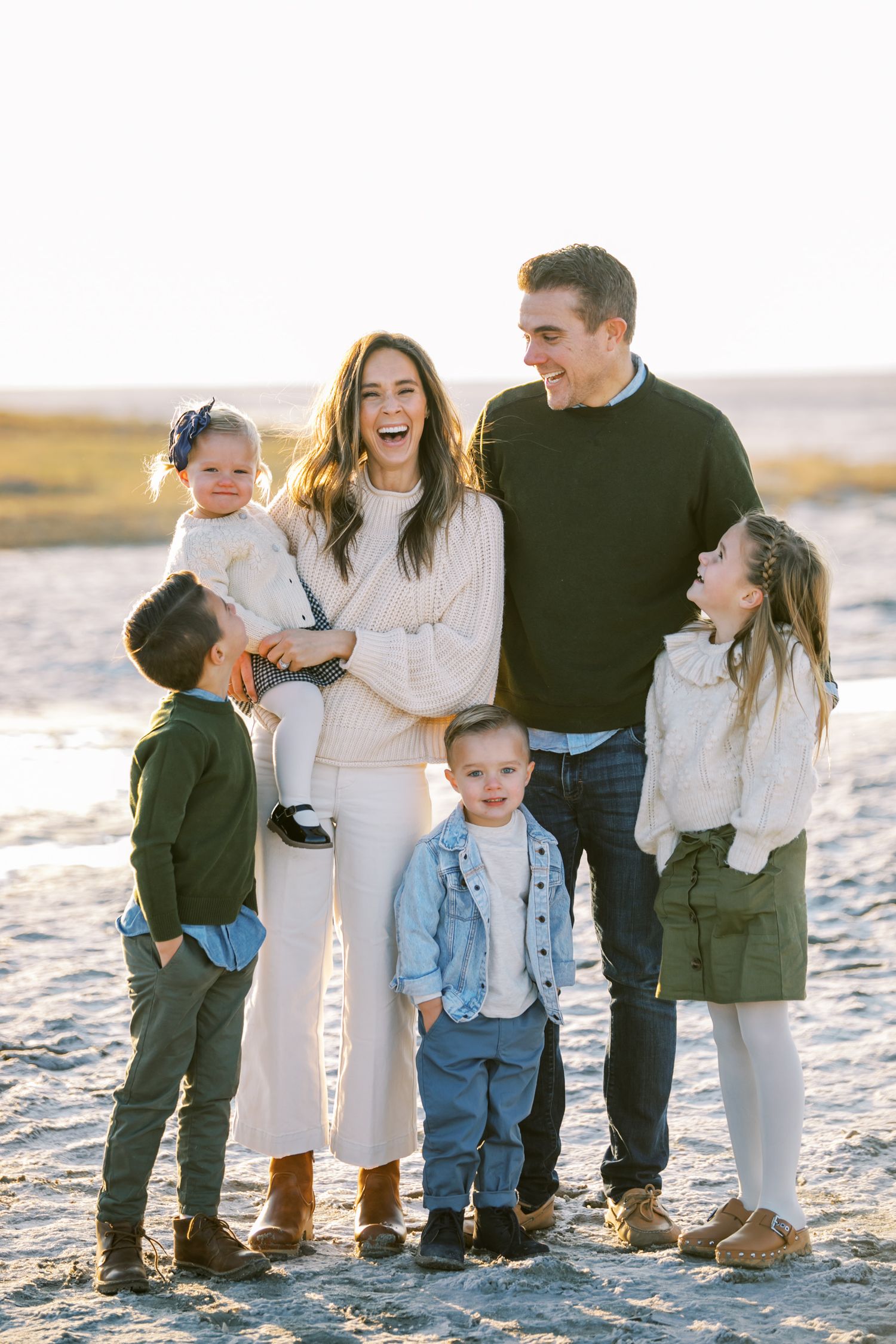 A family poses together barefoot on a sunlit beach in white and dark colored coordinating outfits.
