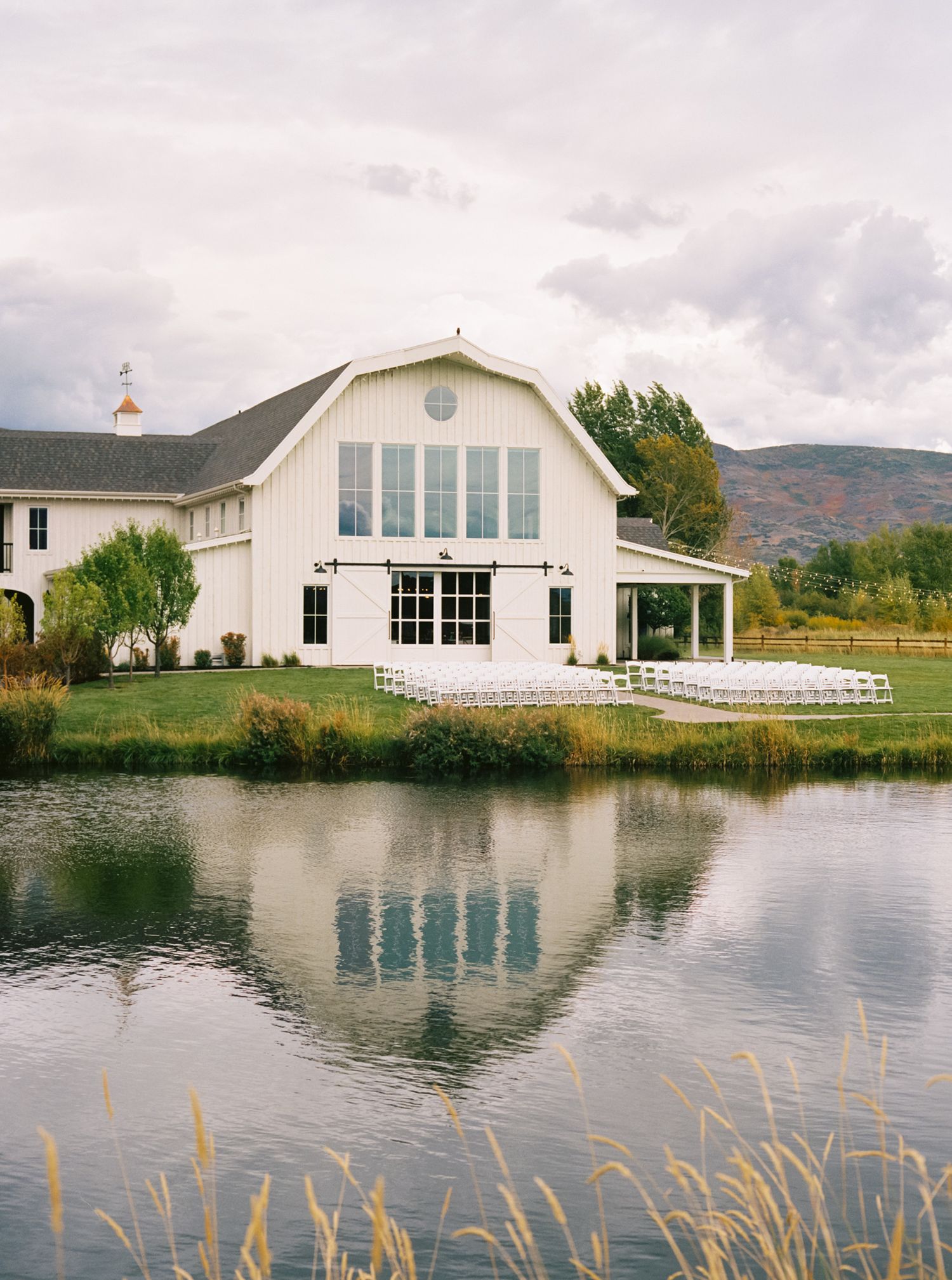 White barn-style wedding venue reflected in a serene pond with mountains in the background on a cloudy day.