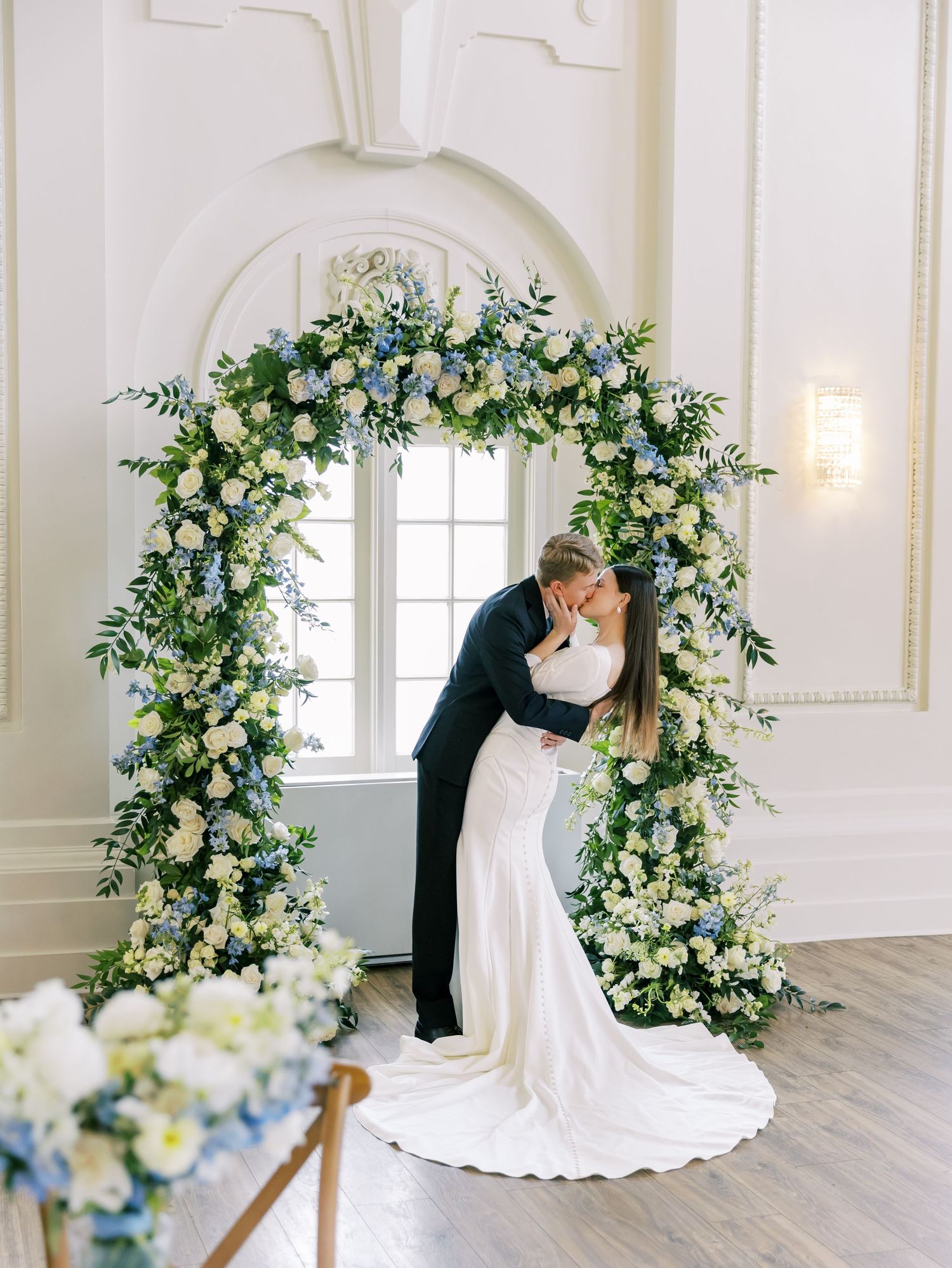 Elegant wedding couple shares intimate moments in a white ballroom decorated with cascading floral arches and greenery.