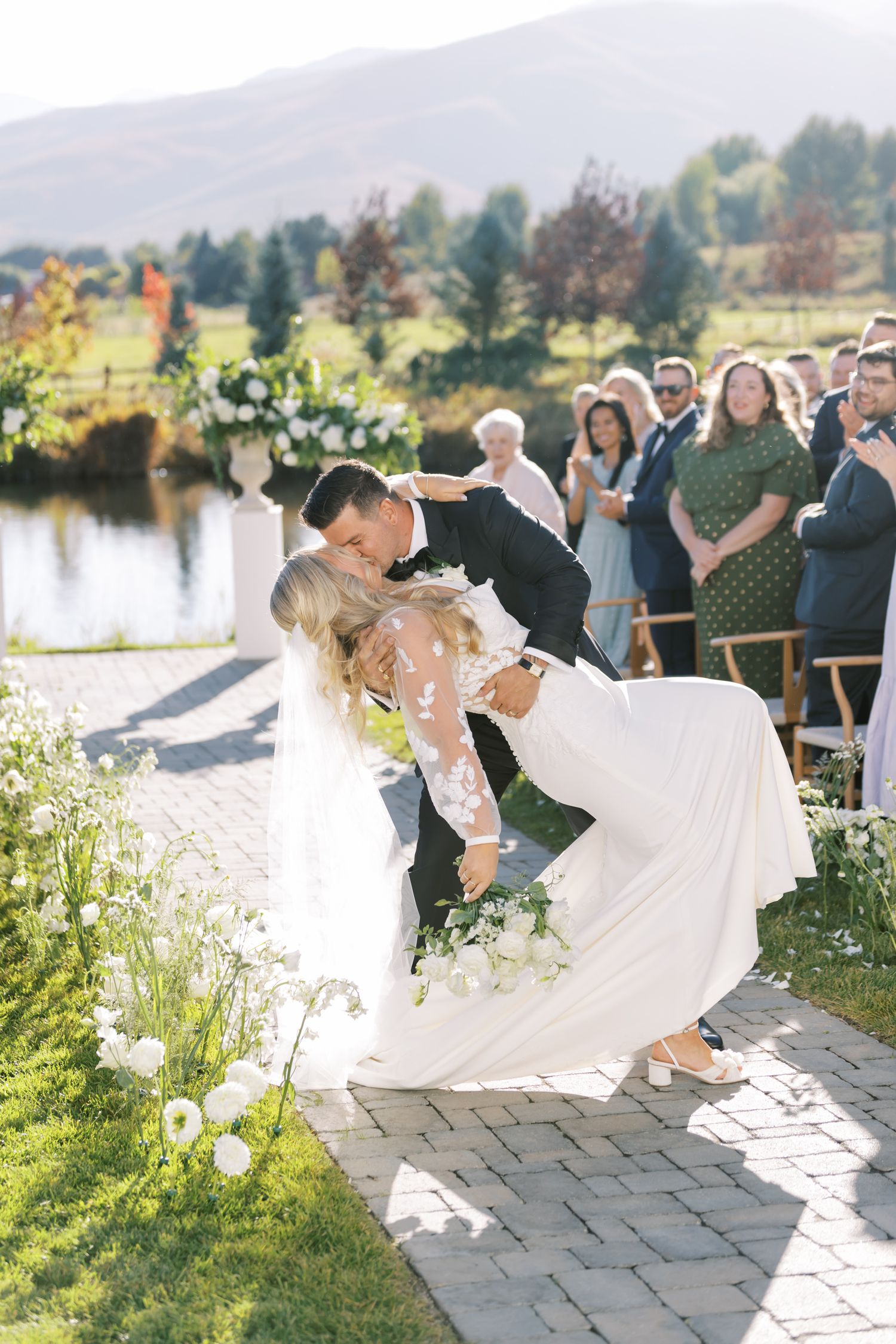 Romantic wedding dip kiss during outdoor ceremony by mountain lake.