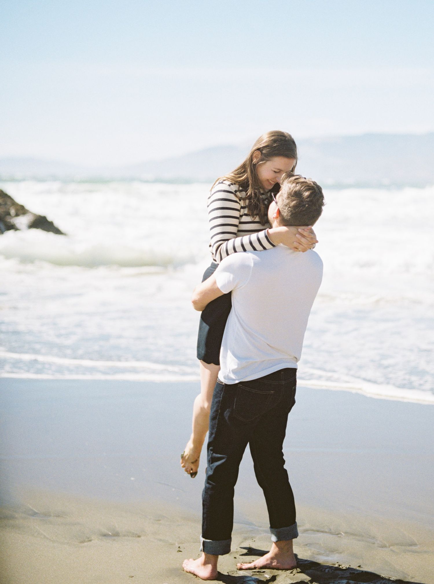 man lifts woman up as they walk barefoot along the san francisco beach