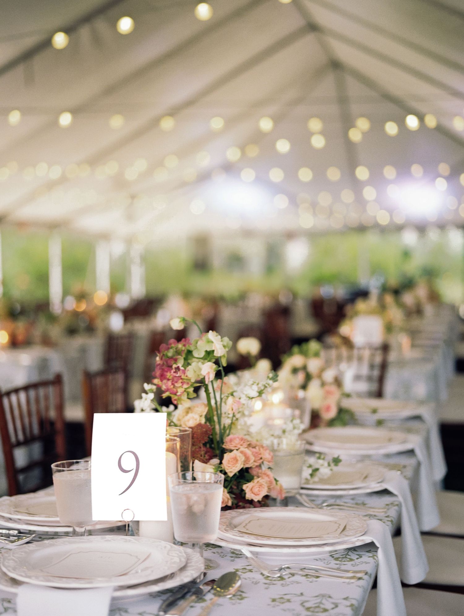 Elegant wedding reception table setting under white tent with string lights and floral centerpieces.