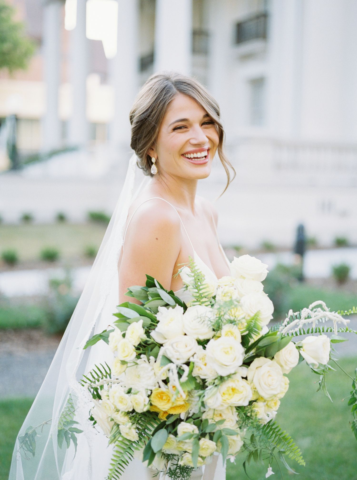 Radiant bride in strapless gown holds lush white and yellow bouquet with greenery while wearing flowing veil outdoors.
