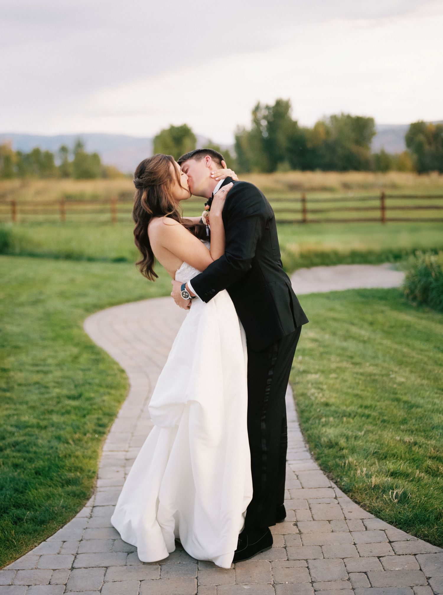 Romantic wedding couple embracing on a stone path surrounded by green fields and mountains.