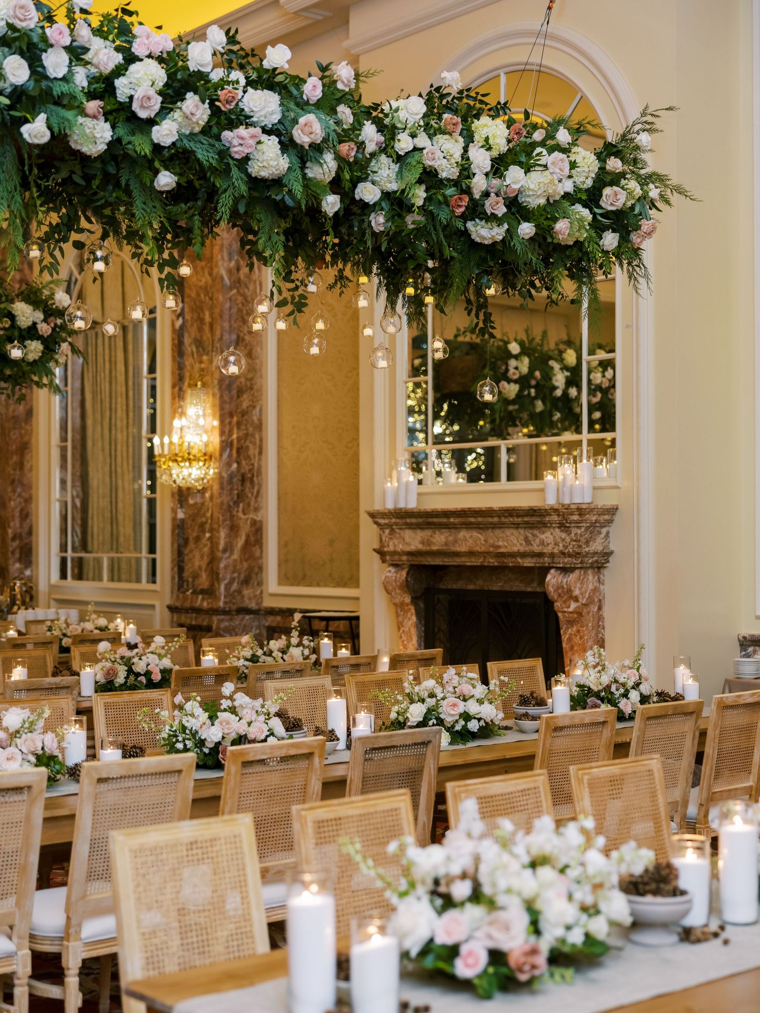 Elegant wedding reception with floral ceiling installation, cane chairs, long tables with candles, and a marble fireplace backdrop.