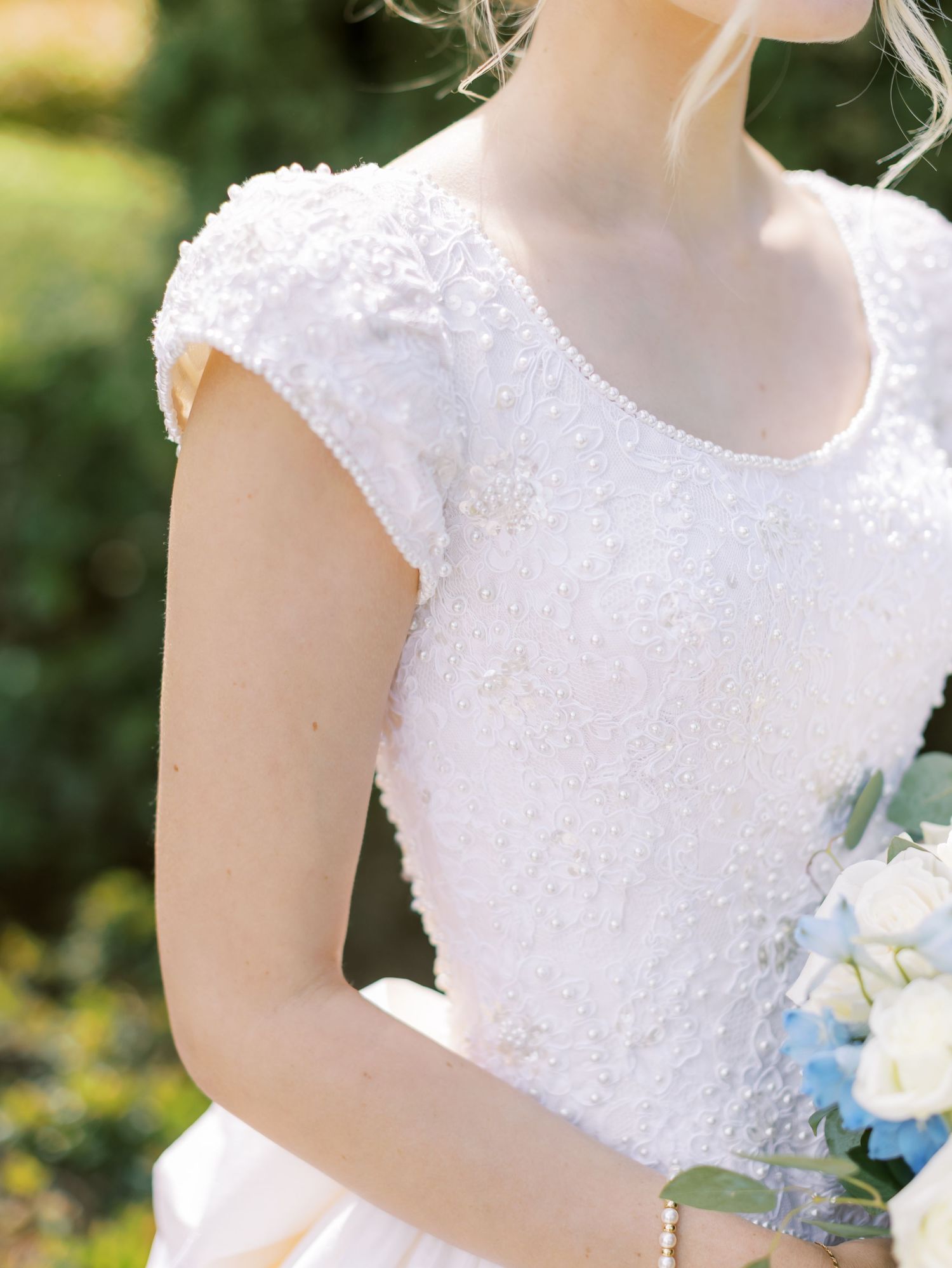 A bride in a white lace wedding dress holds a delicate bouquet with blue and white flowers against a garden backdrop.