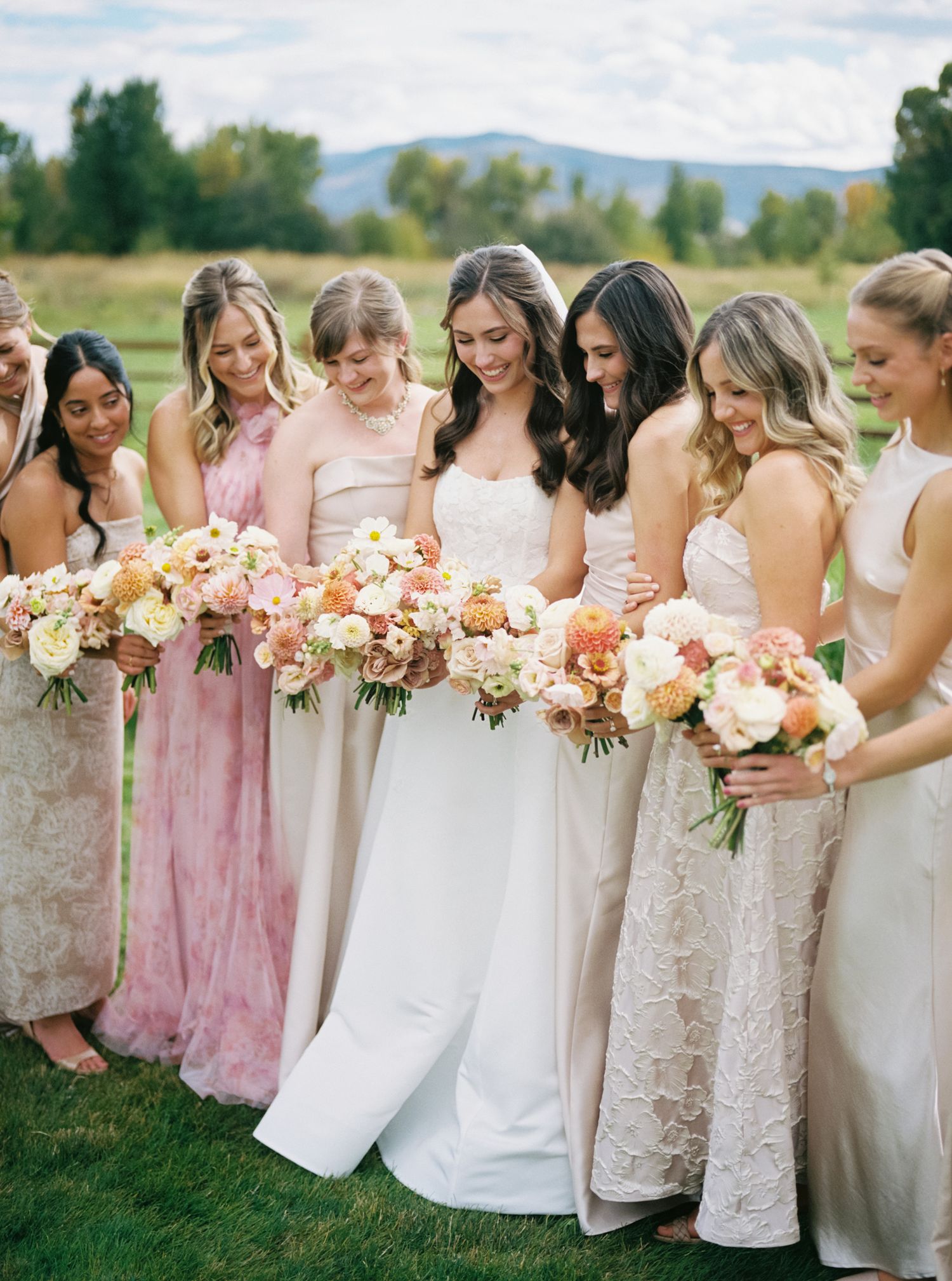 Bridesmaids in blush pink and white dresses hold coordinated peach and coral floral bouquets at an outdoor wedding.