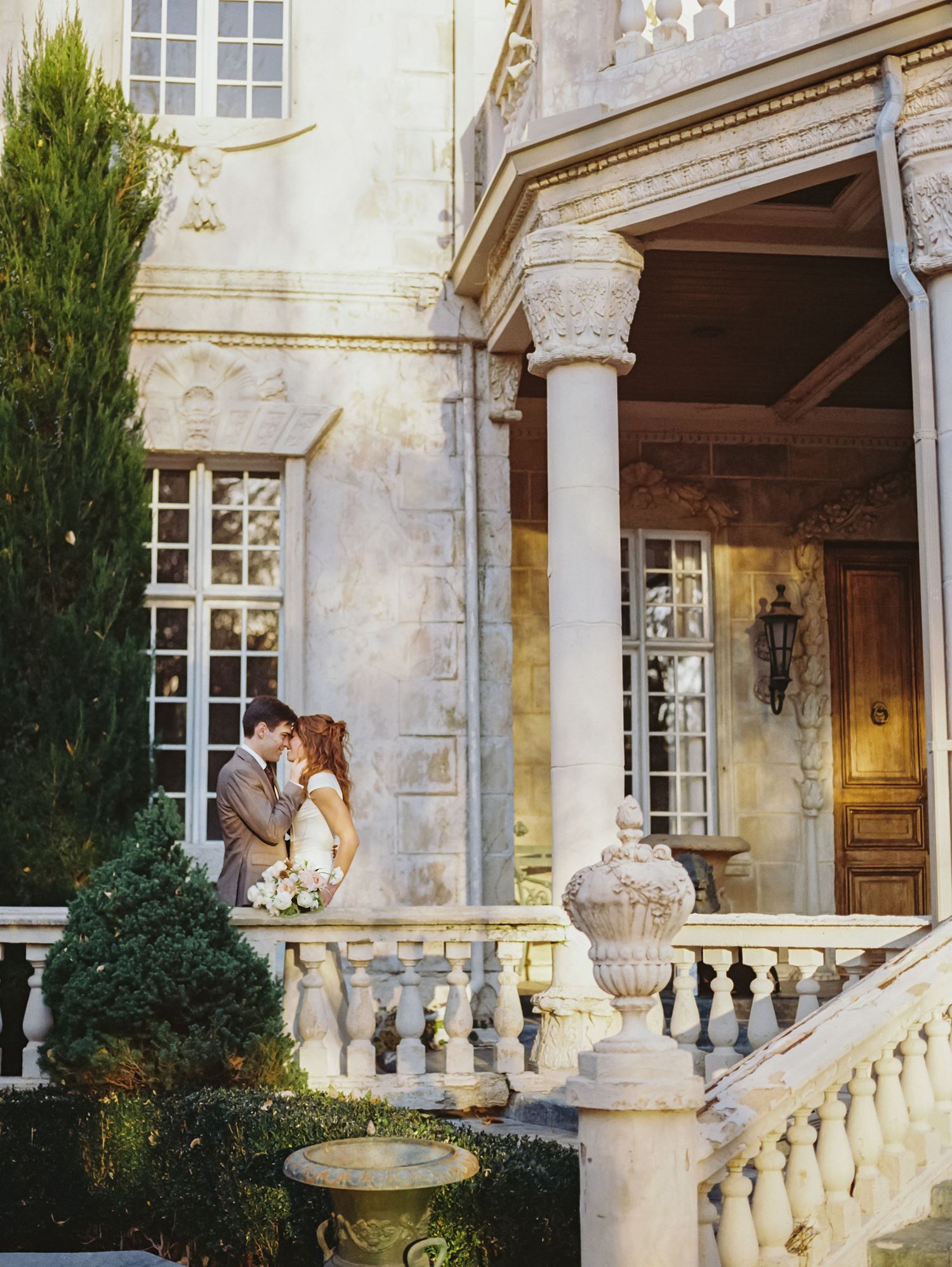 A romantic couple shares a tender moment on an elegant stone balcony with classical architectural details.