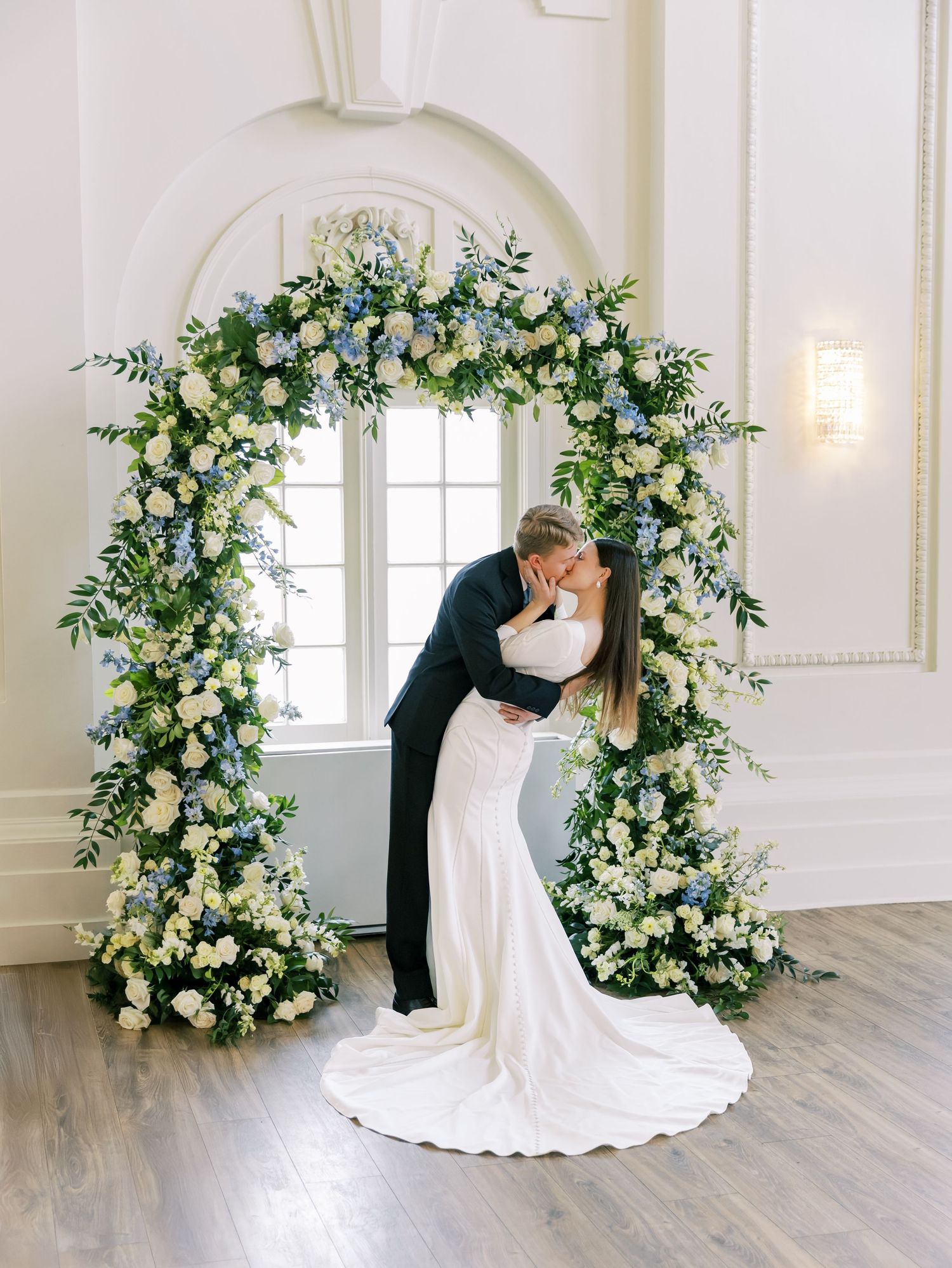 Bride and groom share romantic poses in front of elegant white floral arches in a bright ballroom setting.