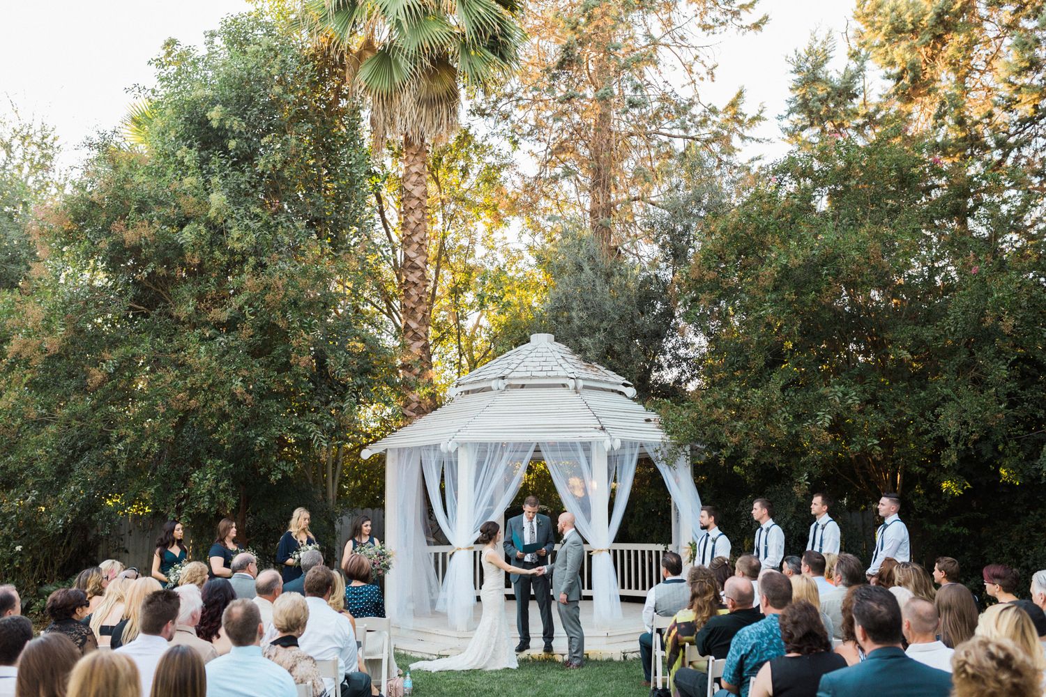 Outdoor wedding ceremony taking place at a white gazebo surrounded by trees and seated guests during golden hour.