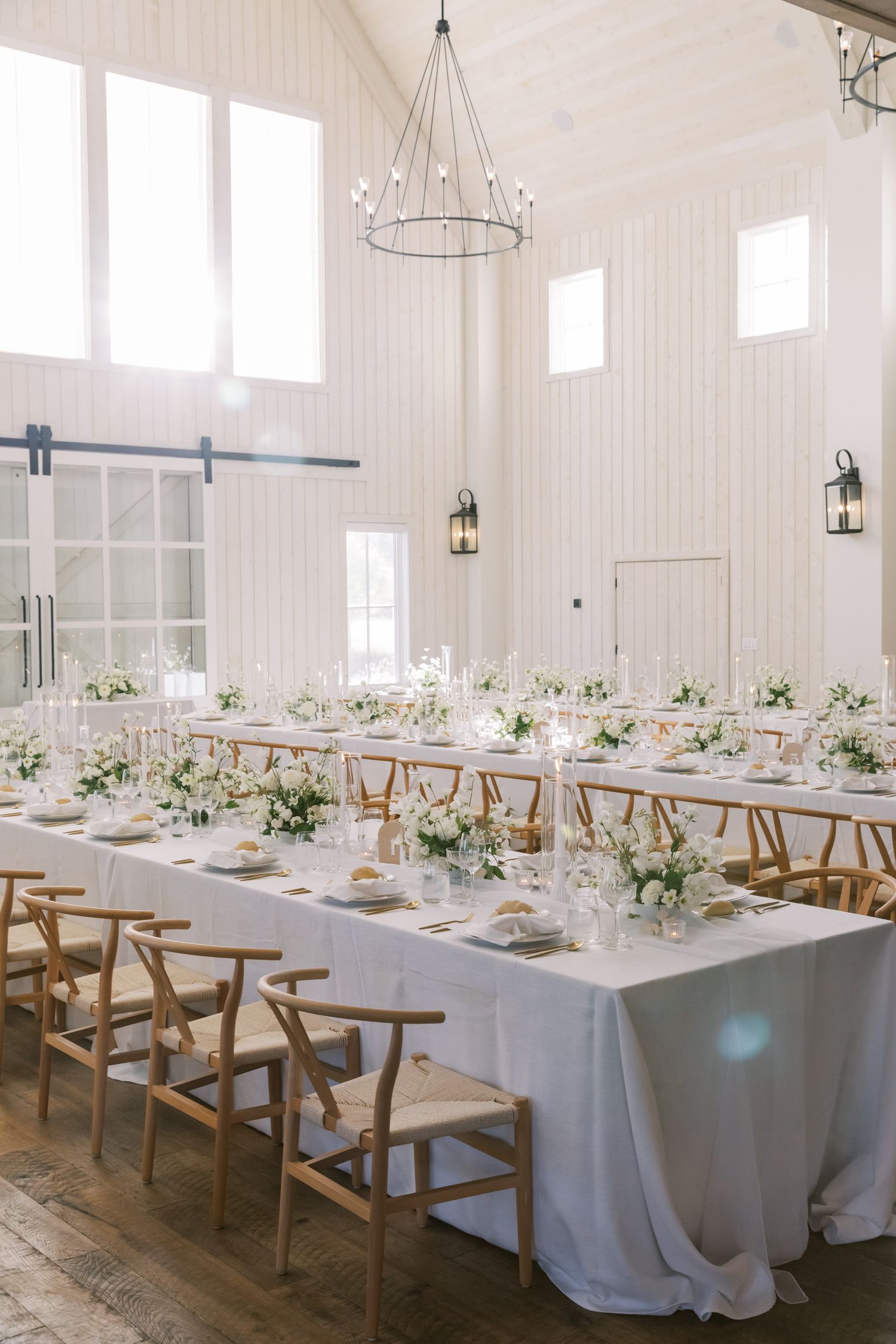 Elegant white wedding reception table setup in a bright barn venue with wooden chairs and chandeliers.