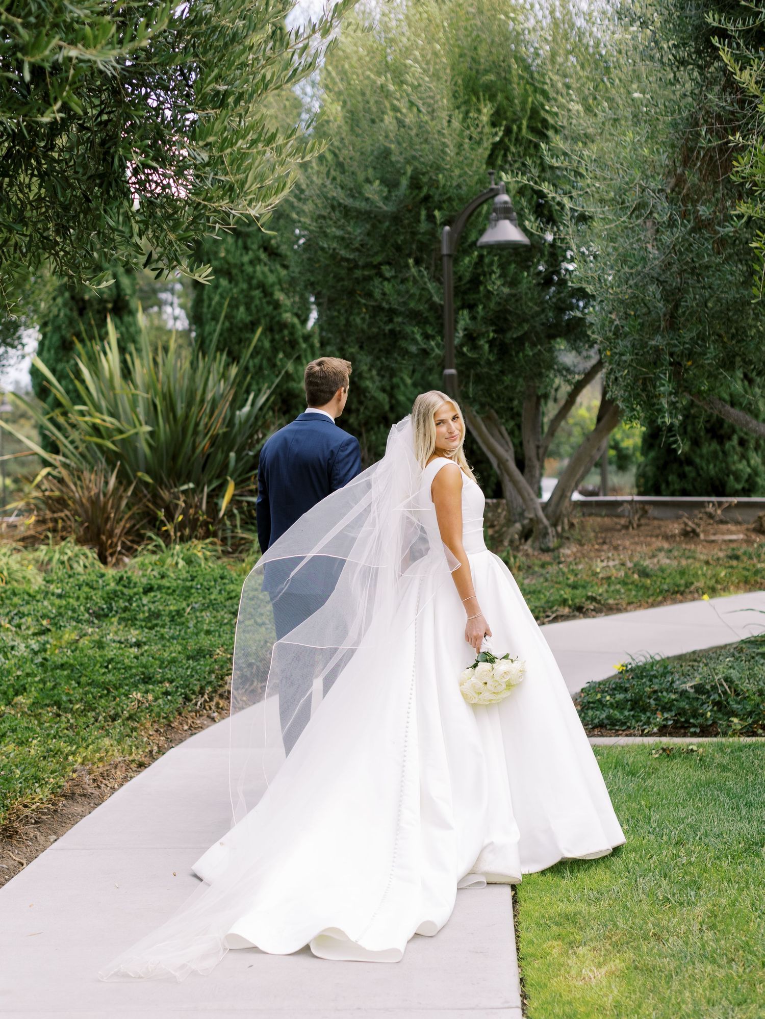 A couple walks along a garden path, with the bride wearing a flowing white gown and long veil.