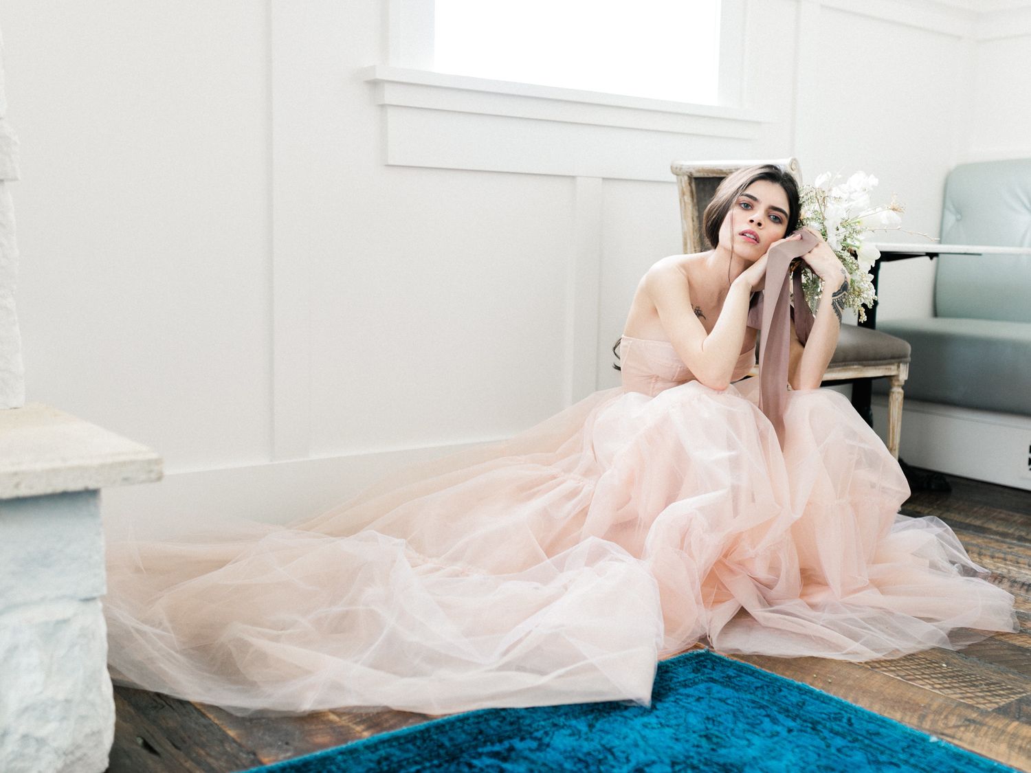 A person in a flowing pink tulle gown poses gracefully on a light gray couch against a modern interior backdrop.