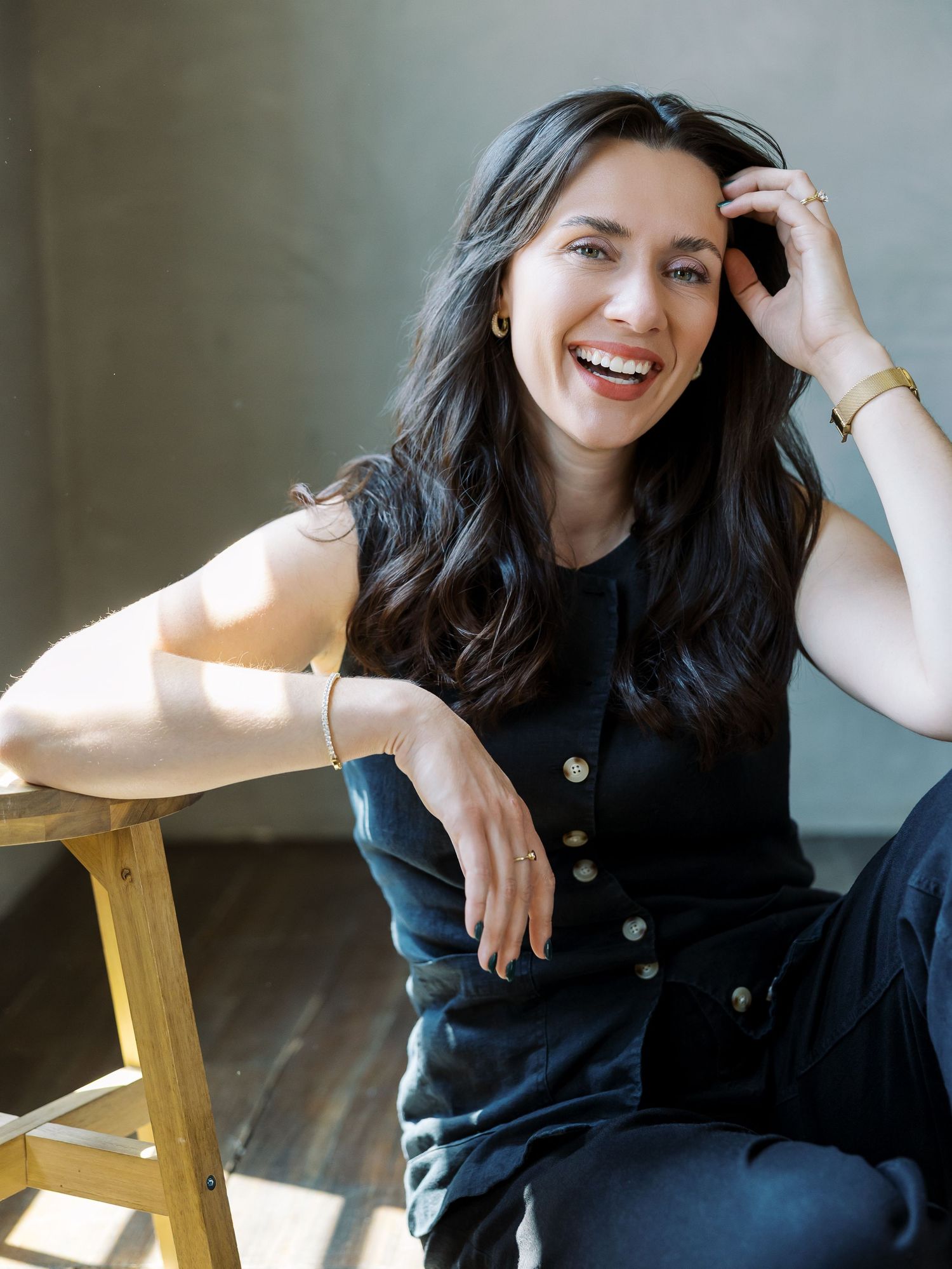 A smiling woman with dark hair sits casually in black outfit, leaning on a wooden stool in a bright studio.