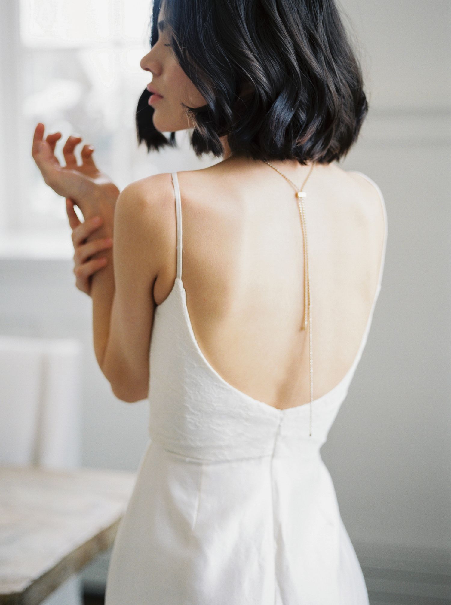 Woman in white low-back wedding dress with gold chain detail, dark bob haircut, elegant bridal fashion.