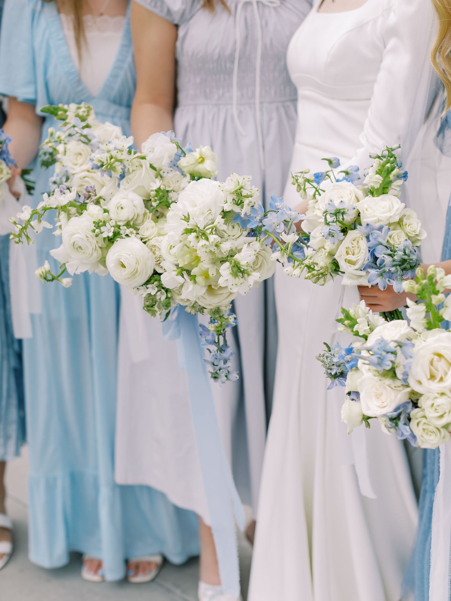 Wedding party members in light blue bridesmaid dresses and white gowns hold elegant white and blue floral bouquets.