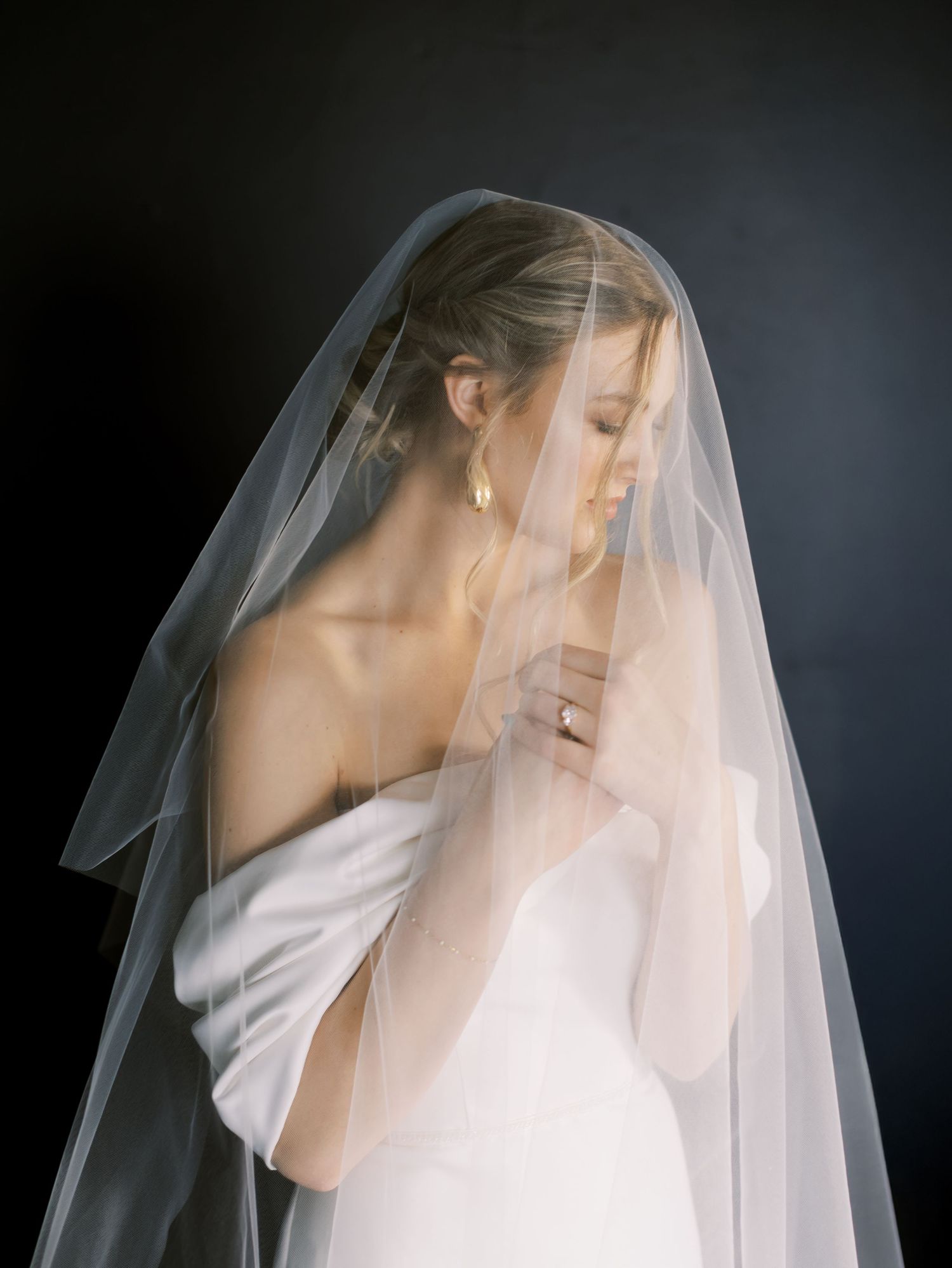 Bride in white strapless gown draped with sheer veil, wearing gold earrings, posed against dark background.