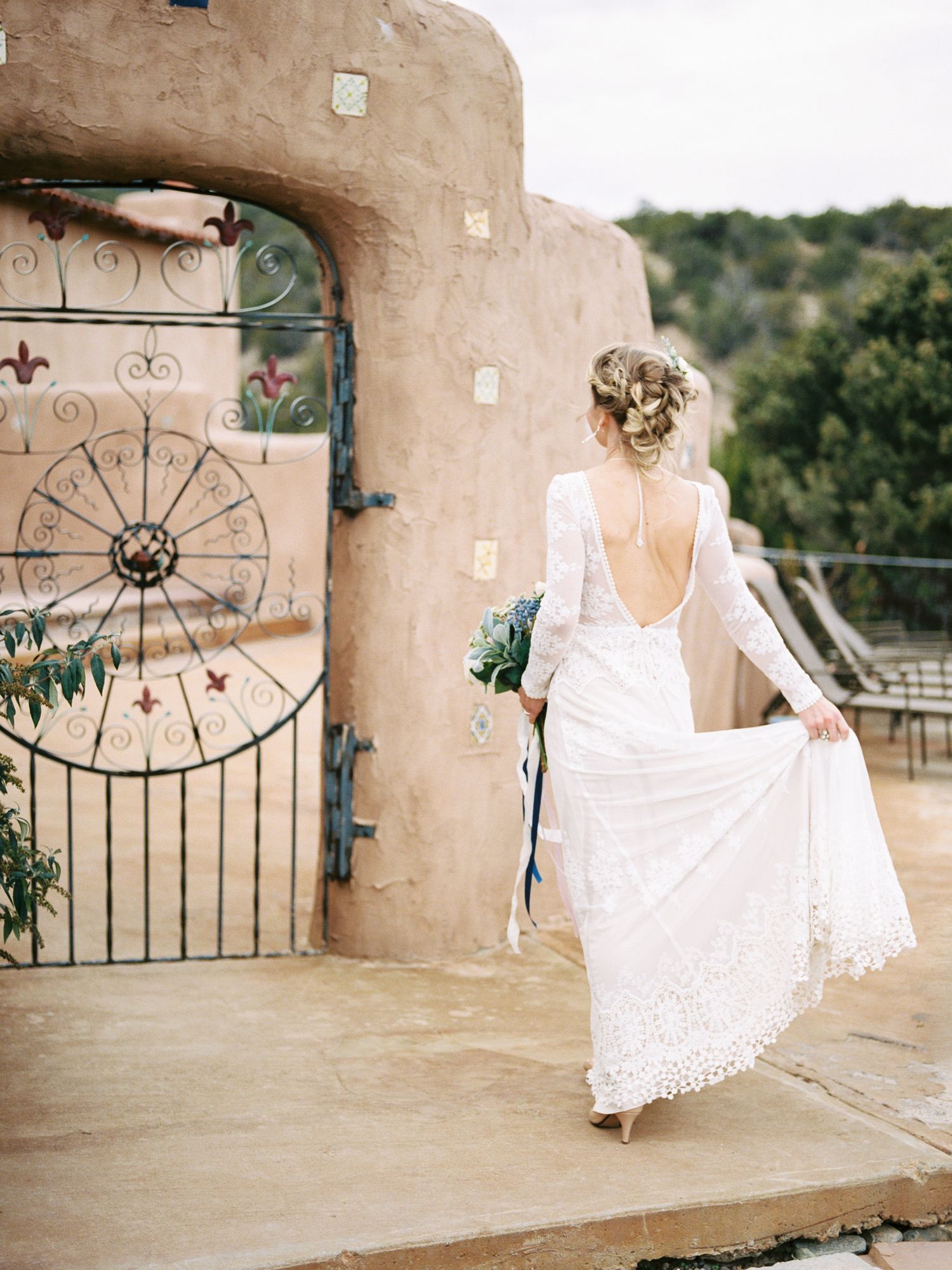 A bride in a white lace backless wedding dress stands by an ornate metal gate at a rustic adobe venue.