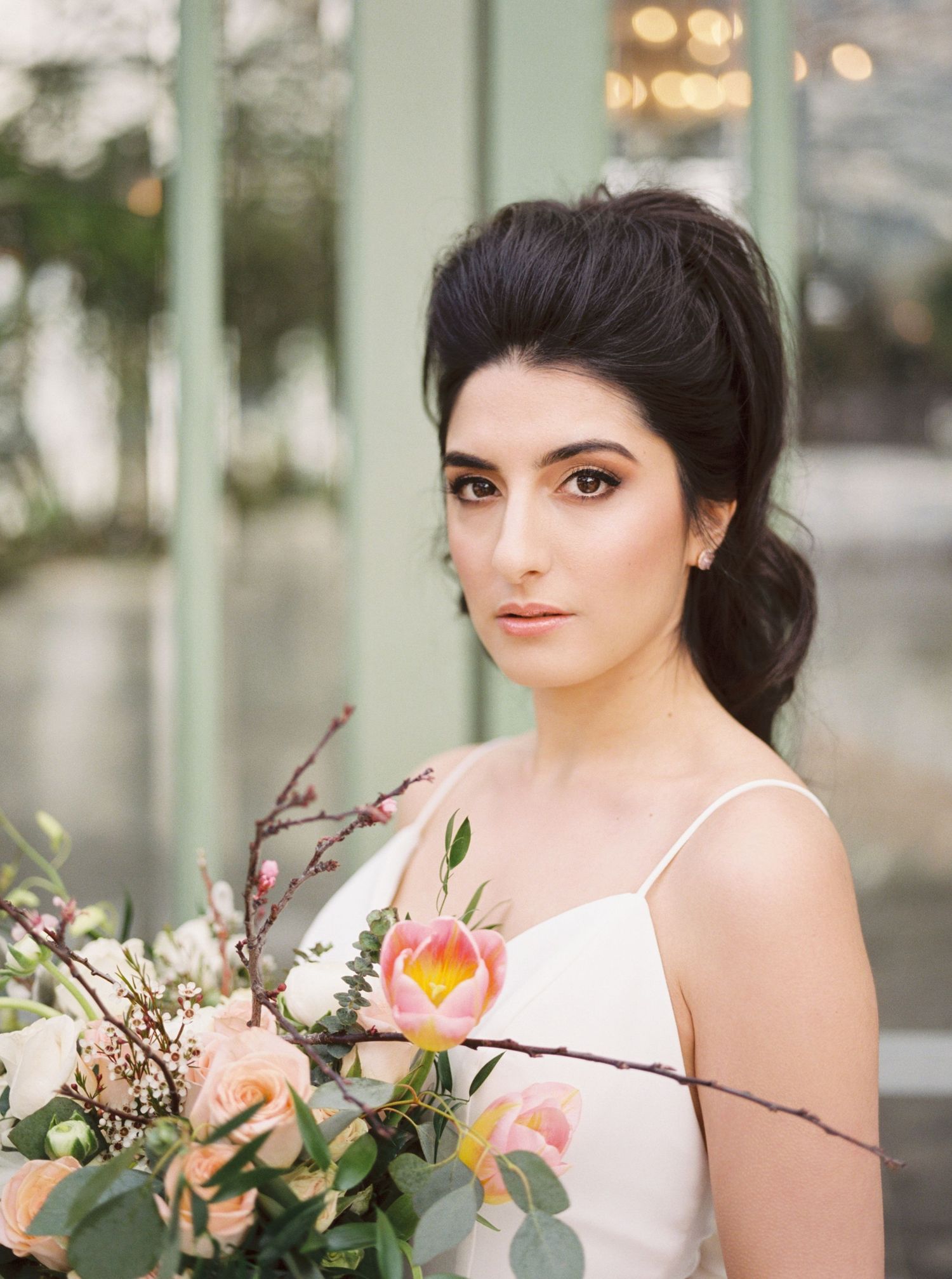 Elegant bride with dark updo holds peach floral bouquet in white gown, posing outdoors near green windows.