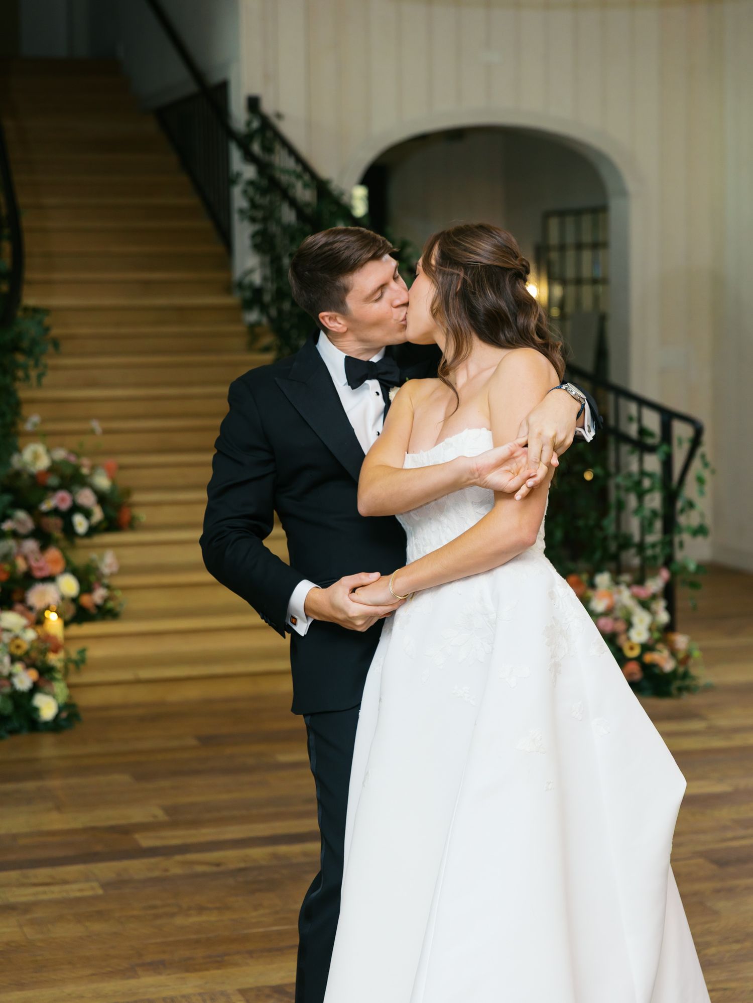 A romantic wedding moment as newlyweds share a kiss on a grand staircase decorated with floral arrangements.