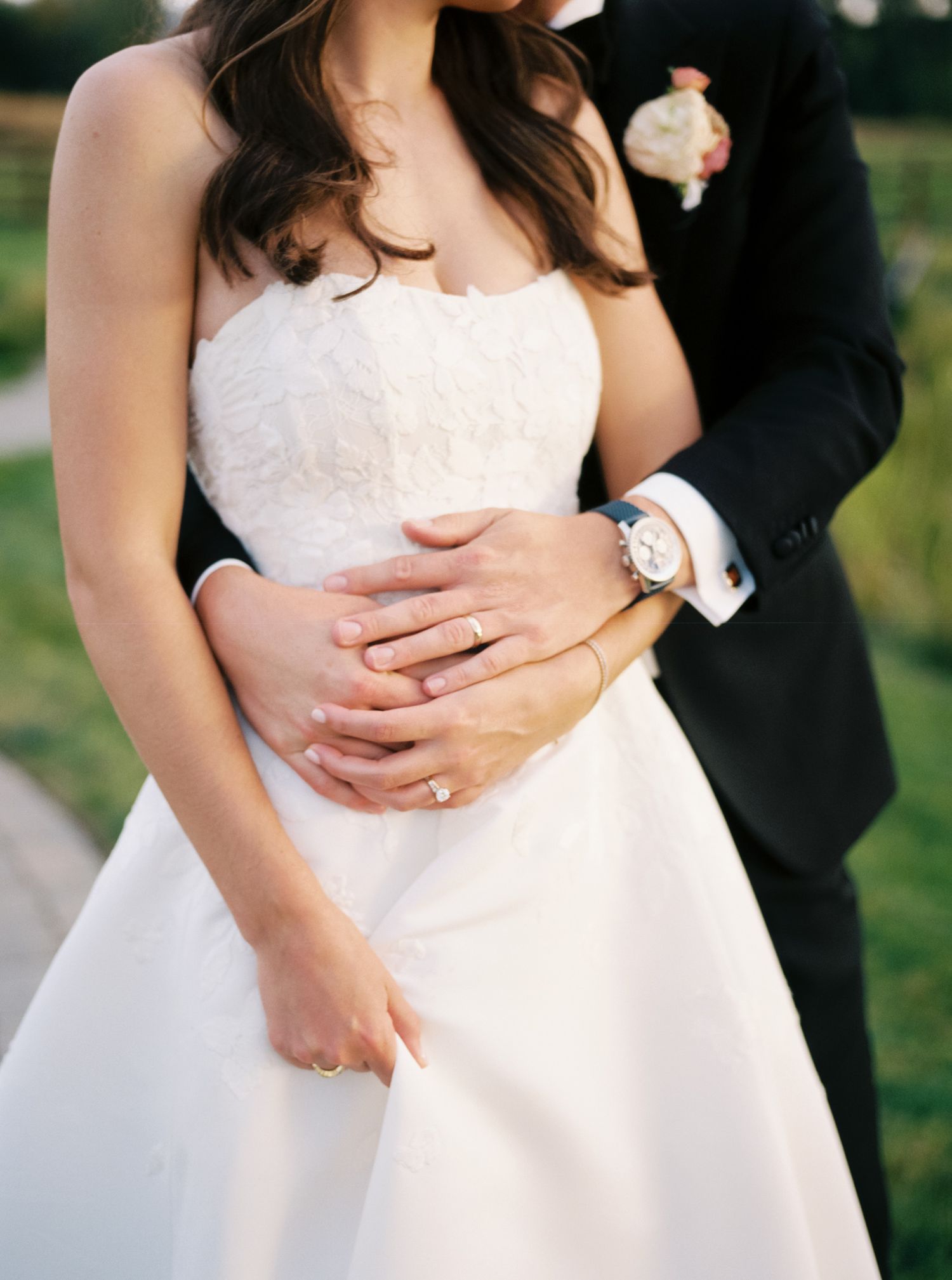 Romantic wedding couple embraces outdoors showing details of white bridal gown and formal black suit.
