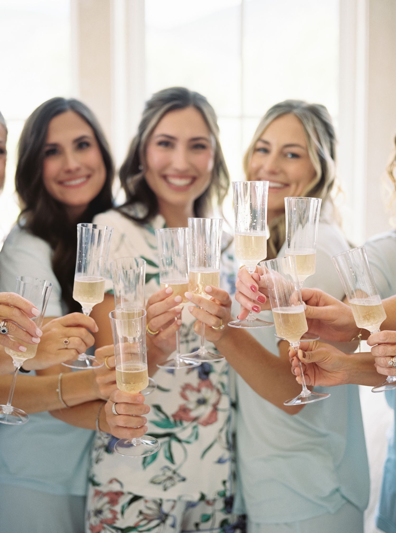 A group toasts with champagne glasses at a celebration wearing floral and light blue dresses.