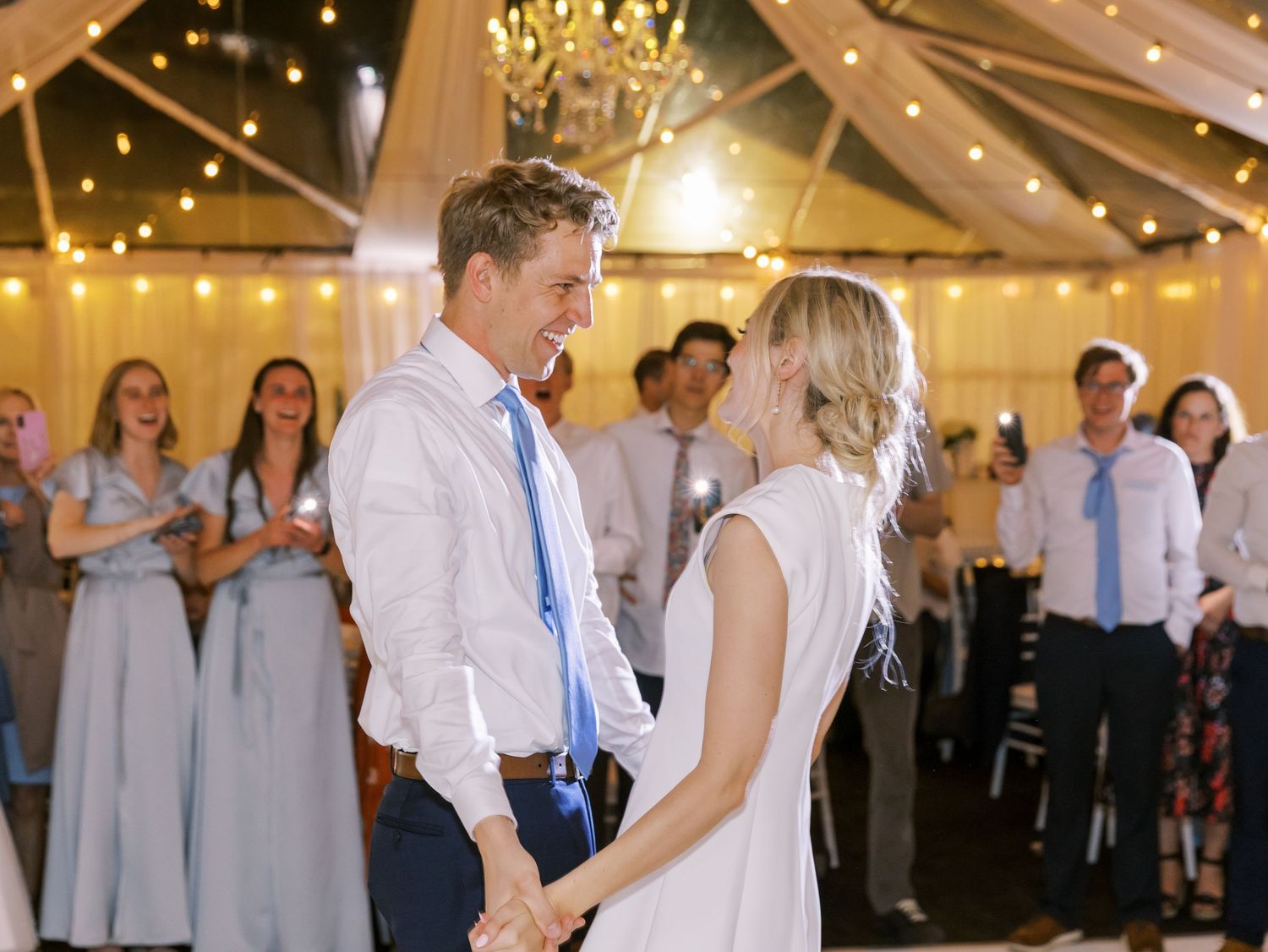 A couple shares their first dance at a wedding reception under twinkling lights and an elegant chandelier.
