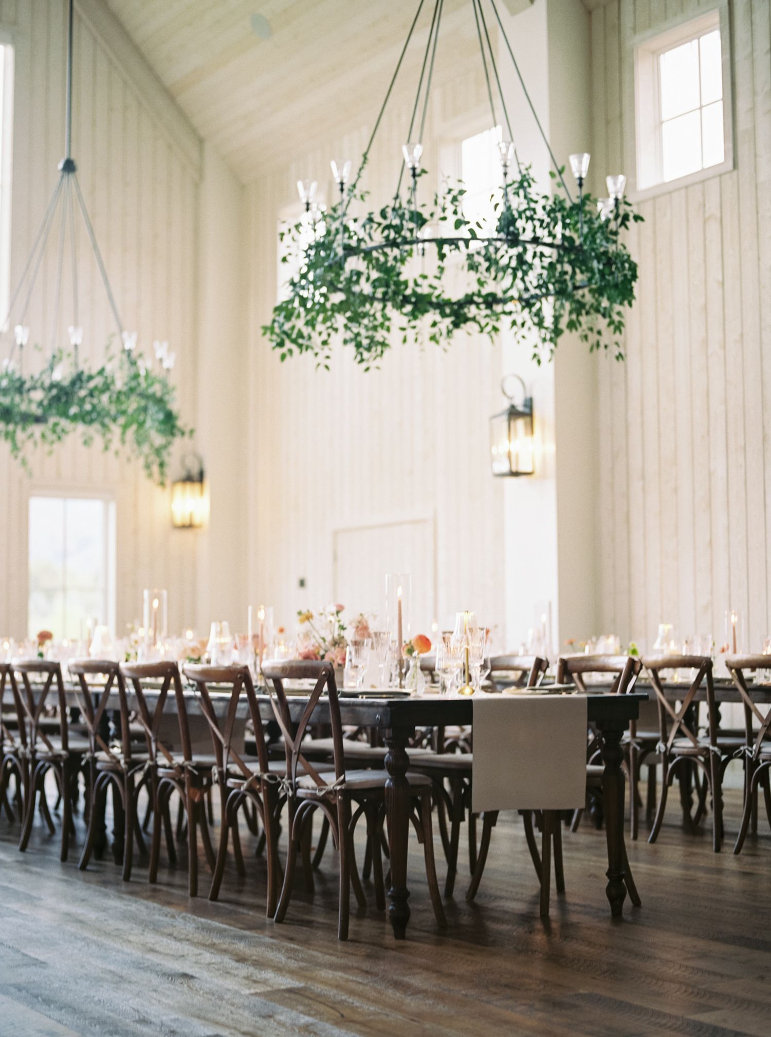 Elegant wedding reception venue with long tables, cross-back chairs, and hanging chandeliers in an all-white room.