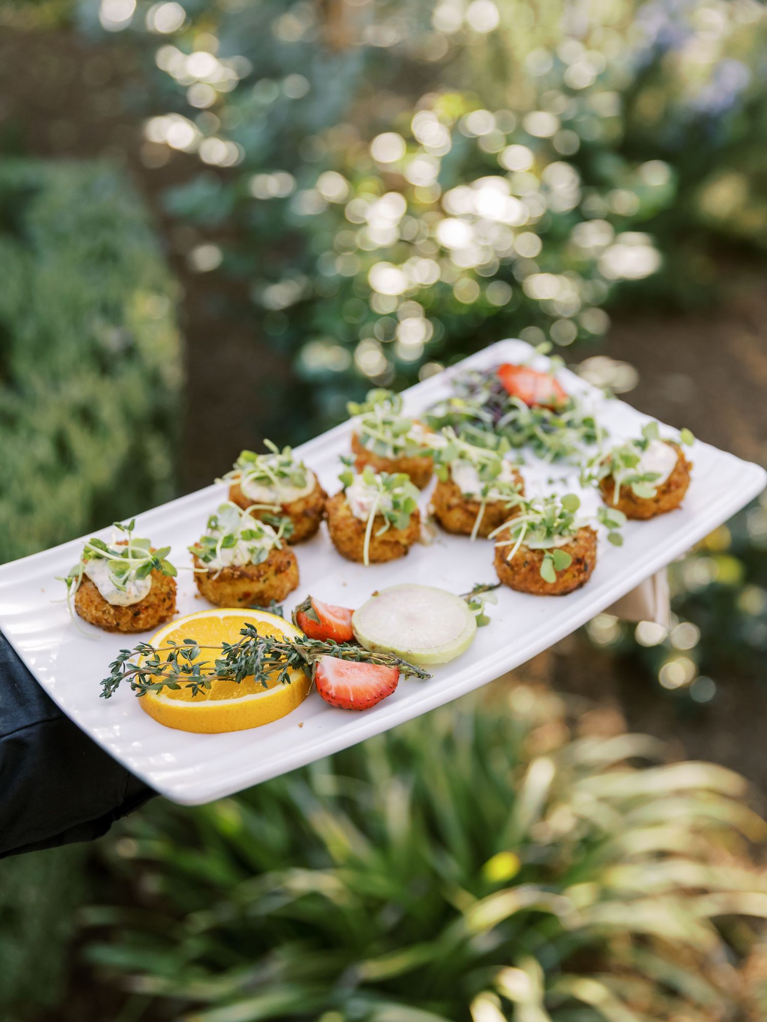 Elegant appetizers on a white platter featuring crostini topped with microgreens and citrus slices in an outdoor garden setting.