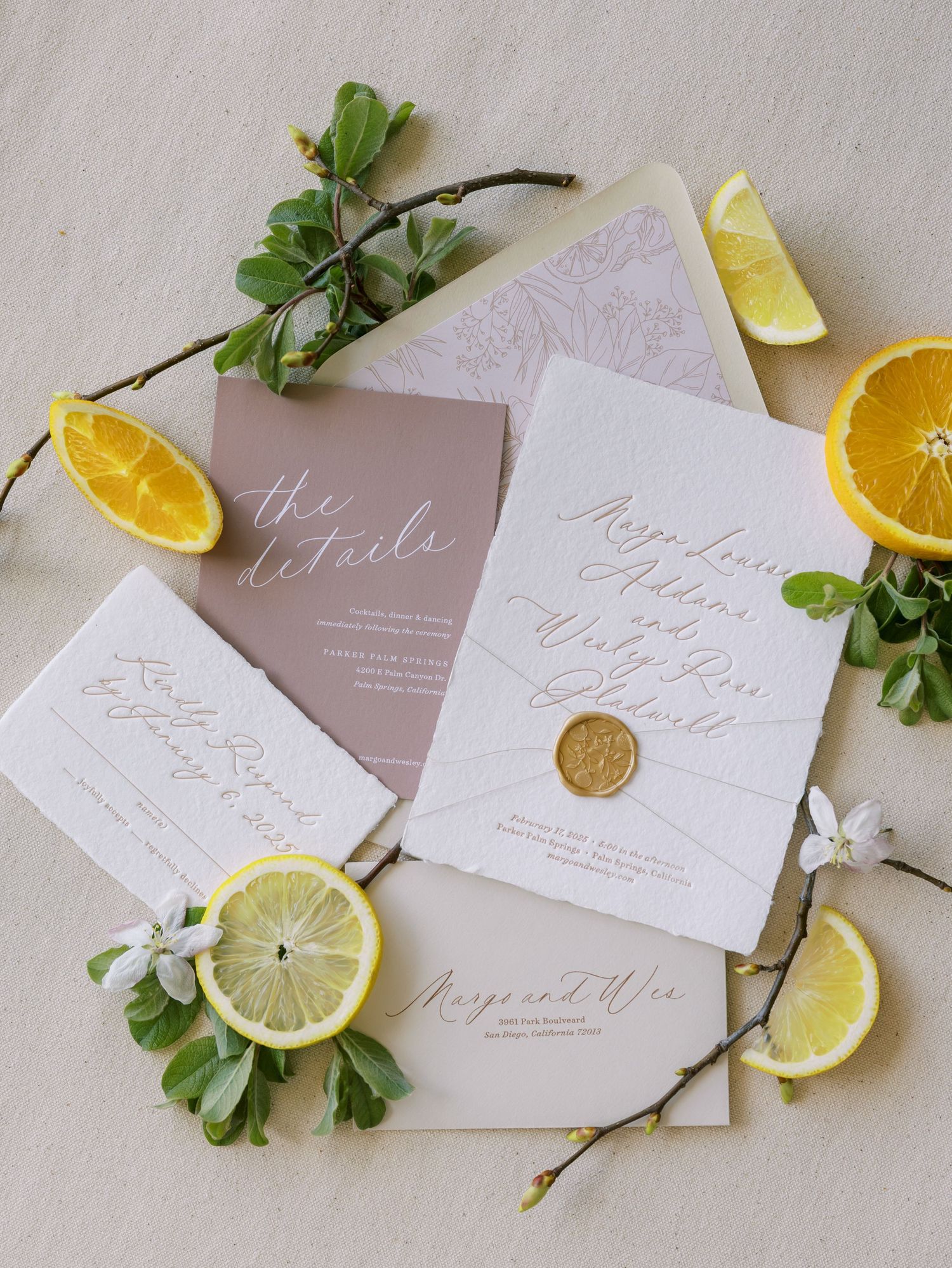 Elegant wedding invitation suite with gold wax seal, mauve envelopes, and calligraphy script, styled with fresh lemon slices and greenery.